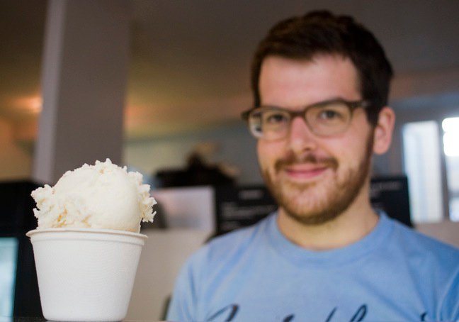 A man with glasses smiles at a scoop of ice cream in a white cup, which is in focus in the foreground, while his background is softly blurred.