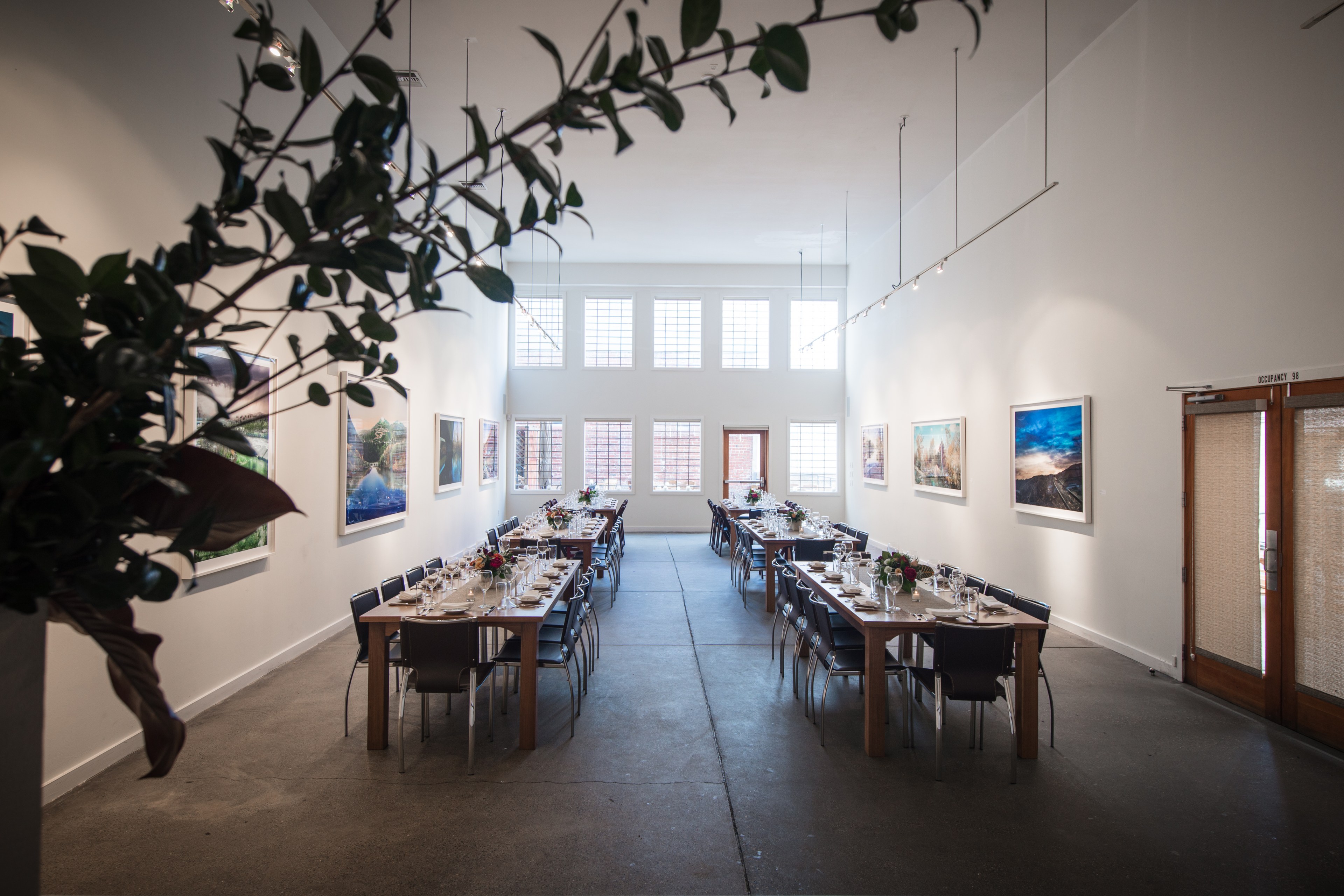 The image shows a long, bright dining room with two tables set for a formal meal, adorned with flowers and surrounded by chairs, with art on the walls.