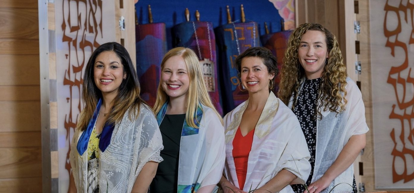 Four women stand side by side, smiling. They are wearing shawls and standing in front of a colorful backdrop with artistic designs and Hebrew letters.