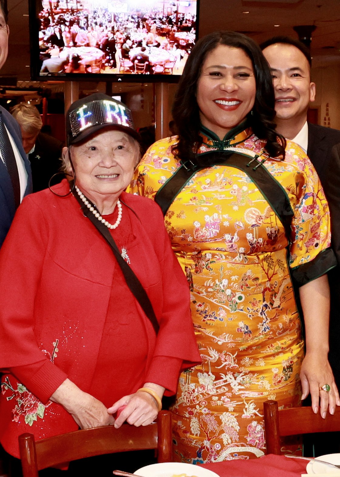 An elderly woman in a red outfit and hat stands beside a smiling woman in a colorful, embroidered dress. There's a crowded event visible in the background.