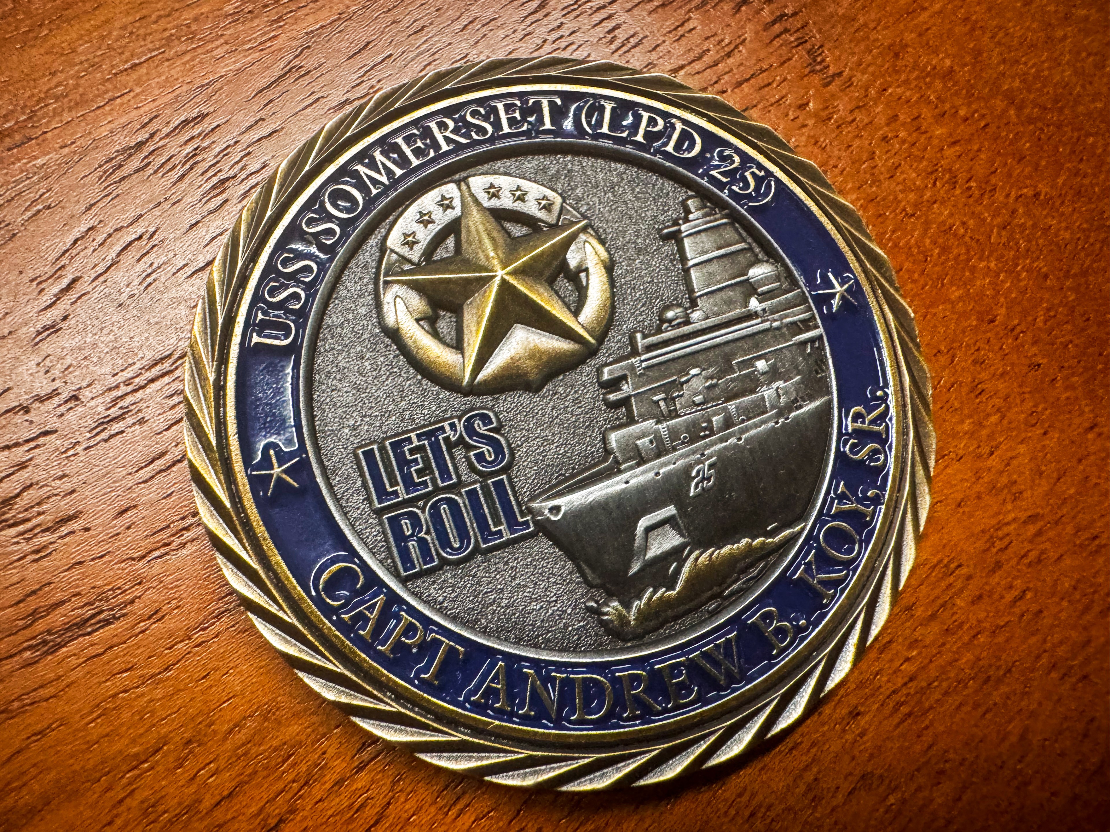 The image shows a round commemorative coin with &quot;USS Somerset (LPD 25)&quot; and &quot;Let's Roll&quot; engraved, featuring a ship and a gold star on a wooden surface.