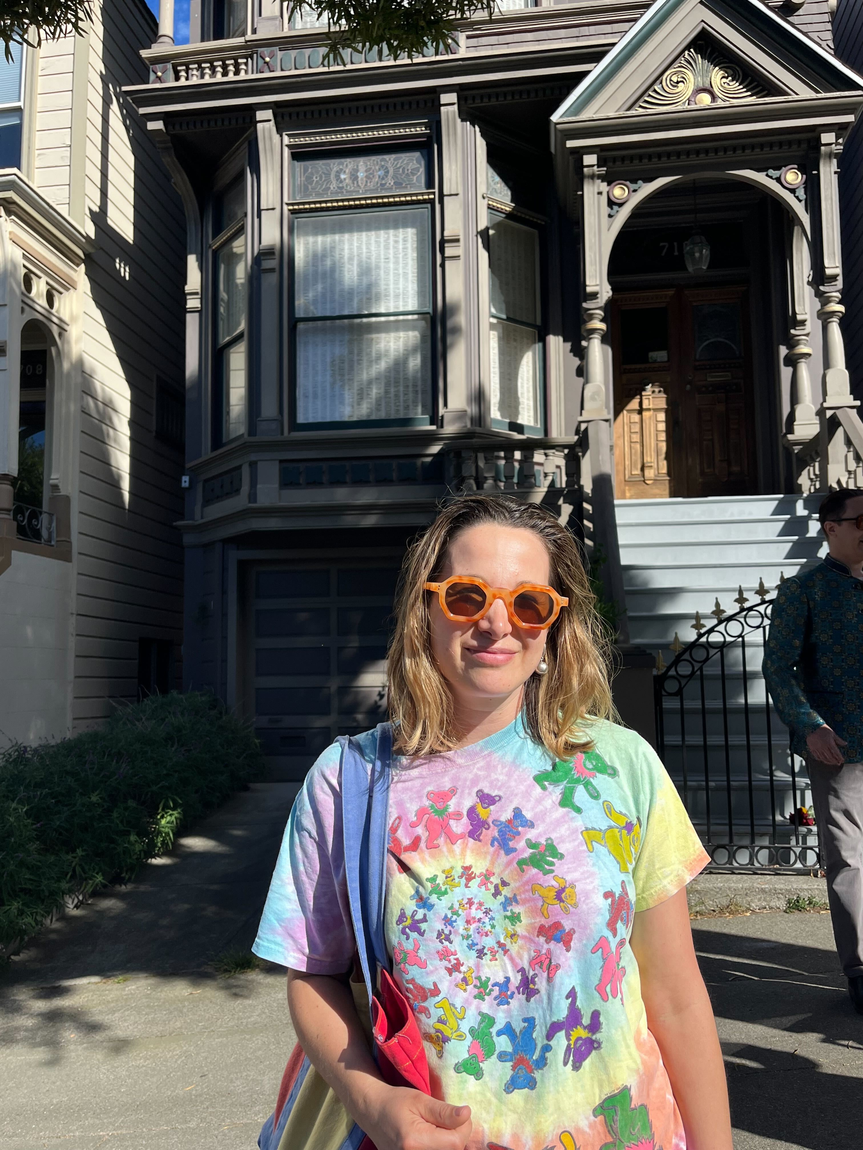 A woman wearing orange sunglasses and a colorful tie-dye shirt stands in front of a Victorian-style house with ornate details and a wooden door.