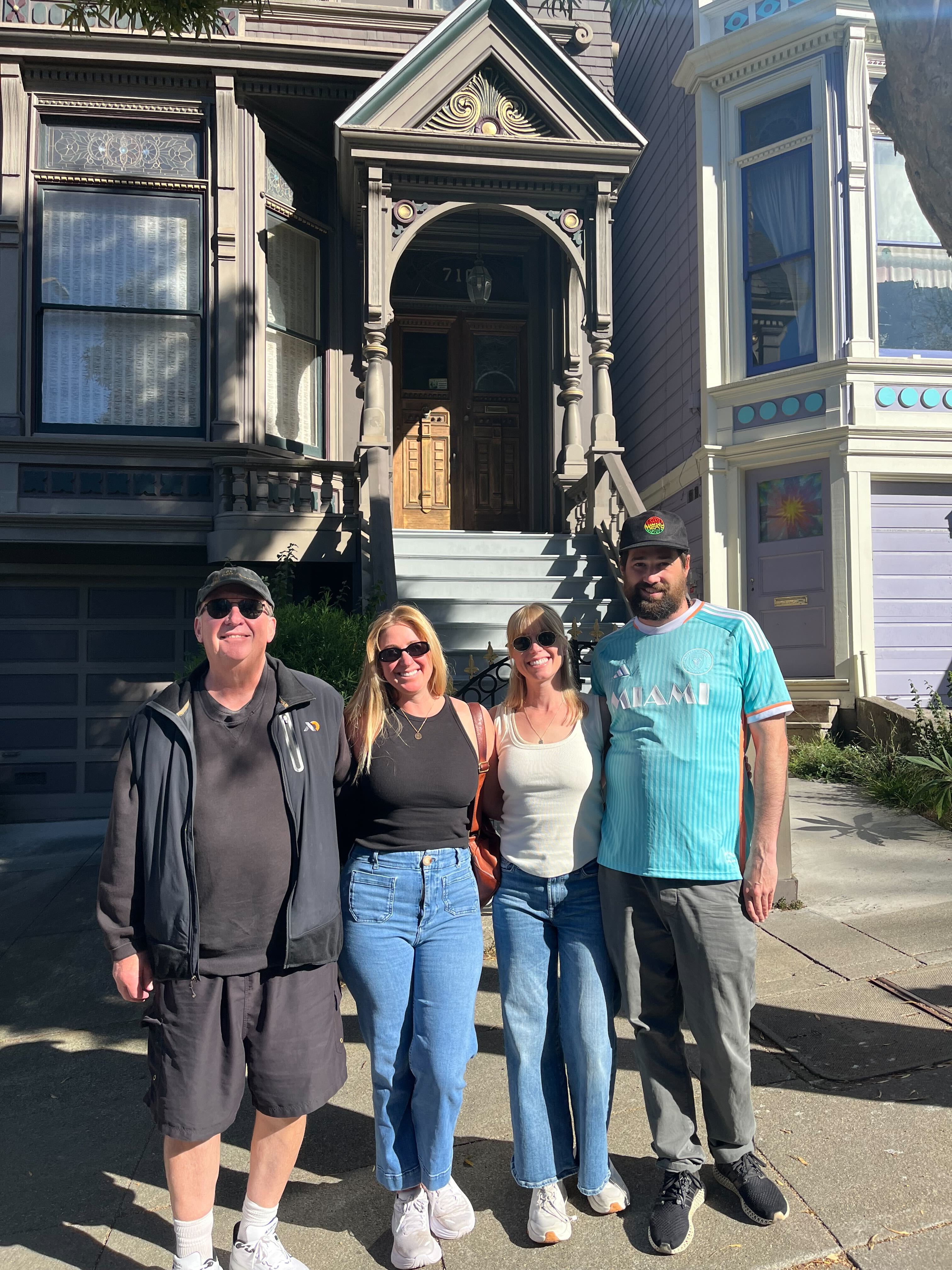 Four people stand smiling in front of a Victorian-style house with intricate details and large windows, under a sunny sky.