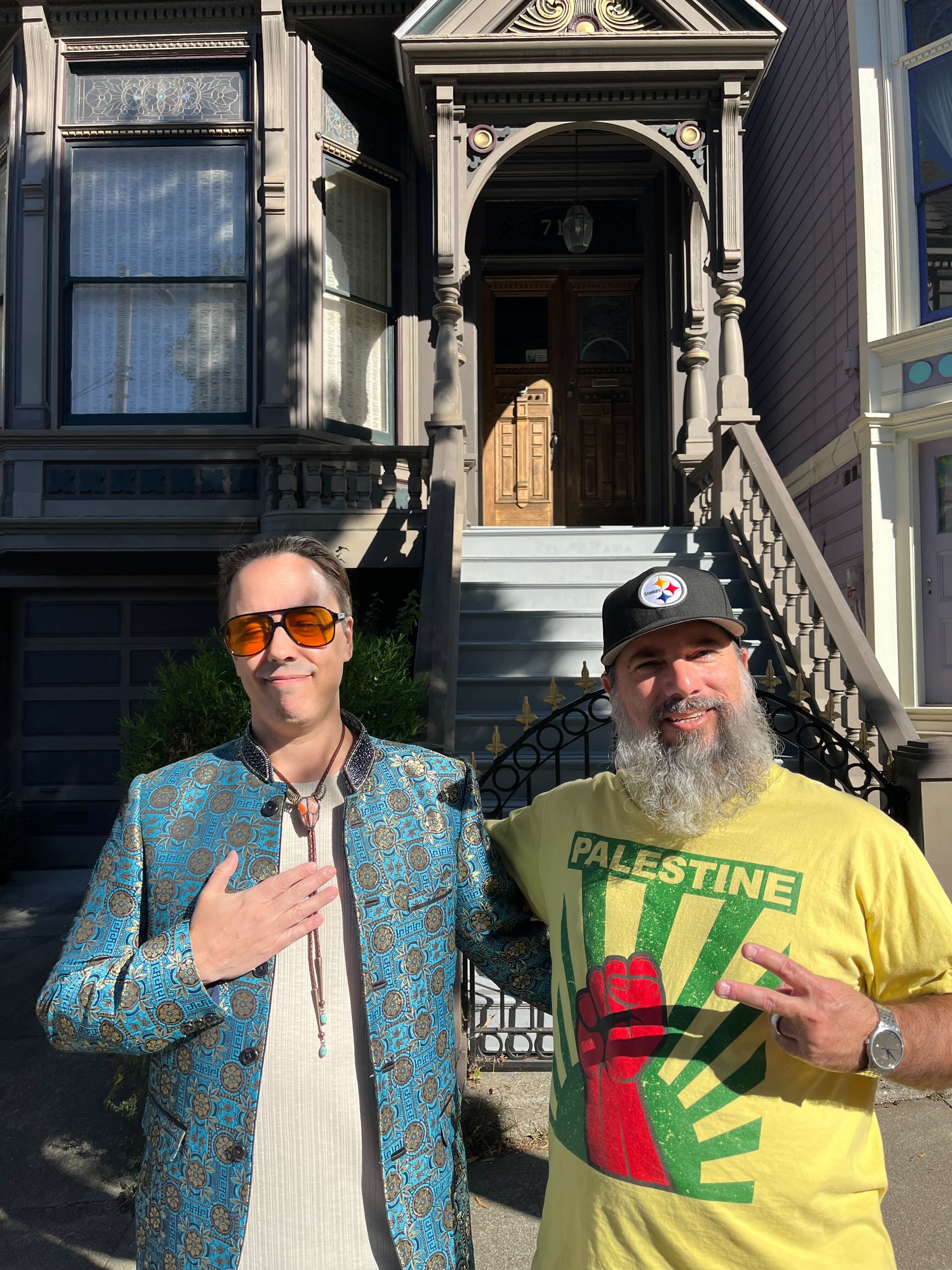 Two men pose in front of a Victorian-style house. The man on the left wears a patterned blue jacket and sunglasses, while the man on the right wears a yellow &quot;Palestine&quot; T-shirt.