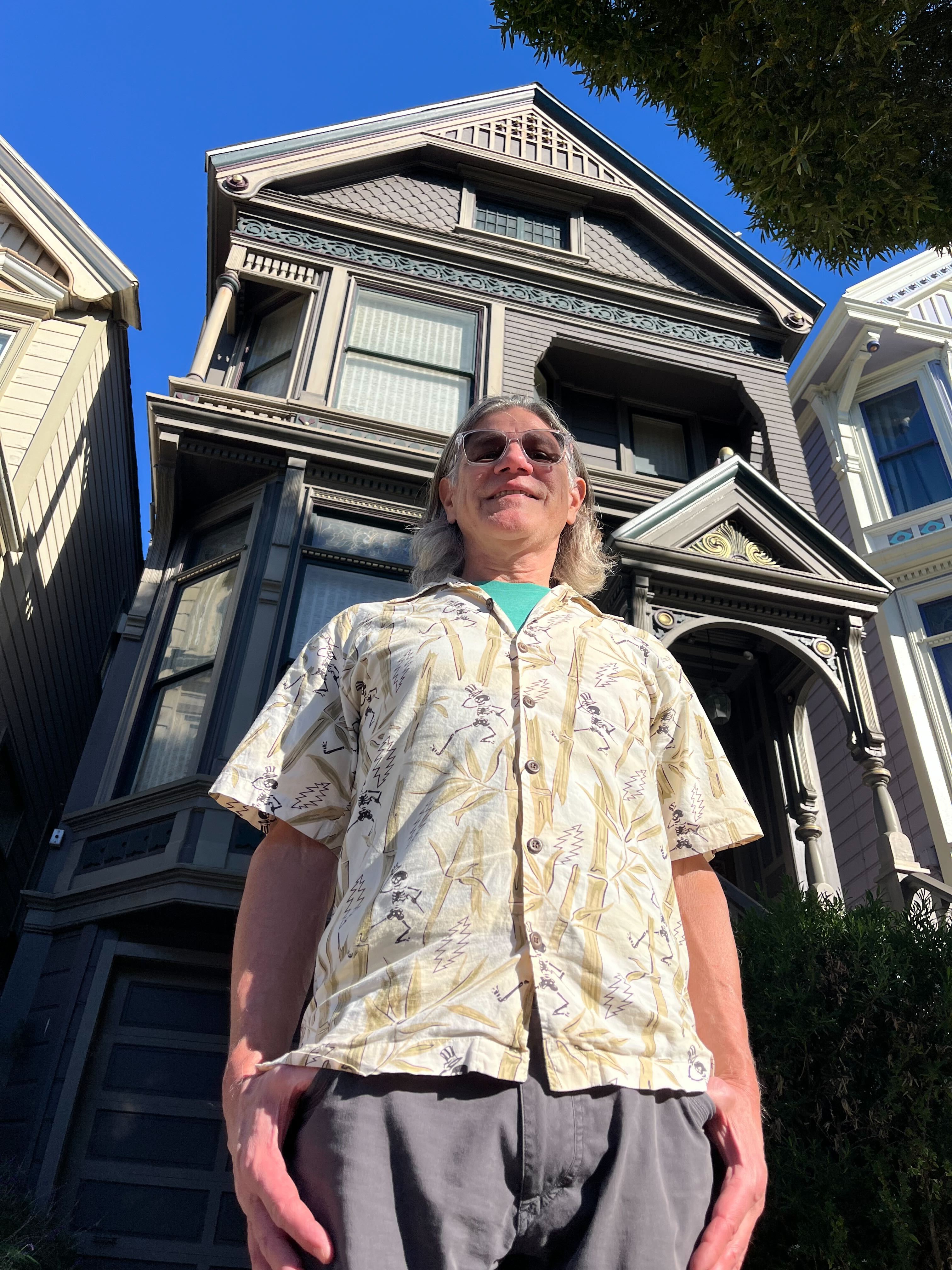 A person in a patterned shirt stands in front of a detailed, multi-story Victorian house with a bright blue sky above.