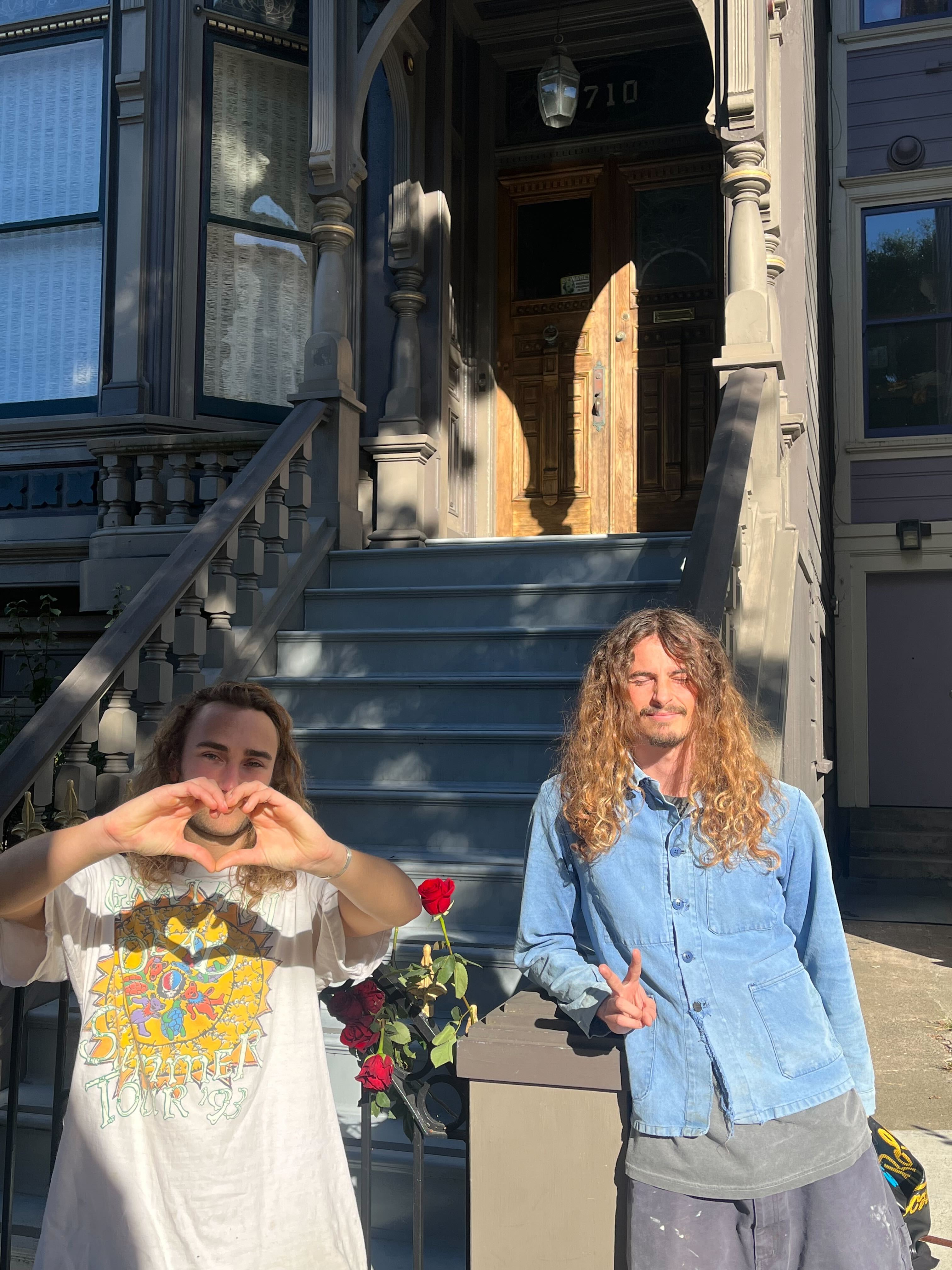 Two people with long hair stand in front of a traditional house. One is making a heart shape with hands, wearing a graphic tee; the other shows a peace sign. Roses are nearby.