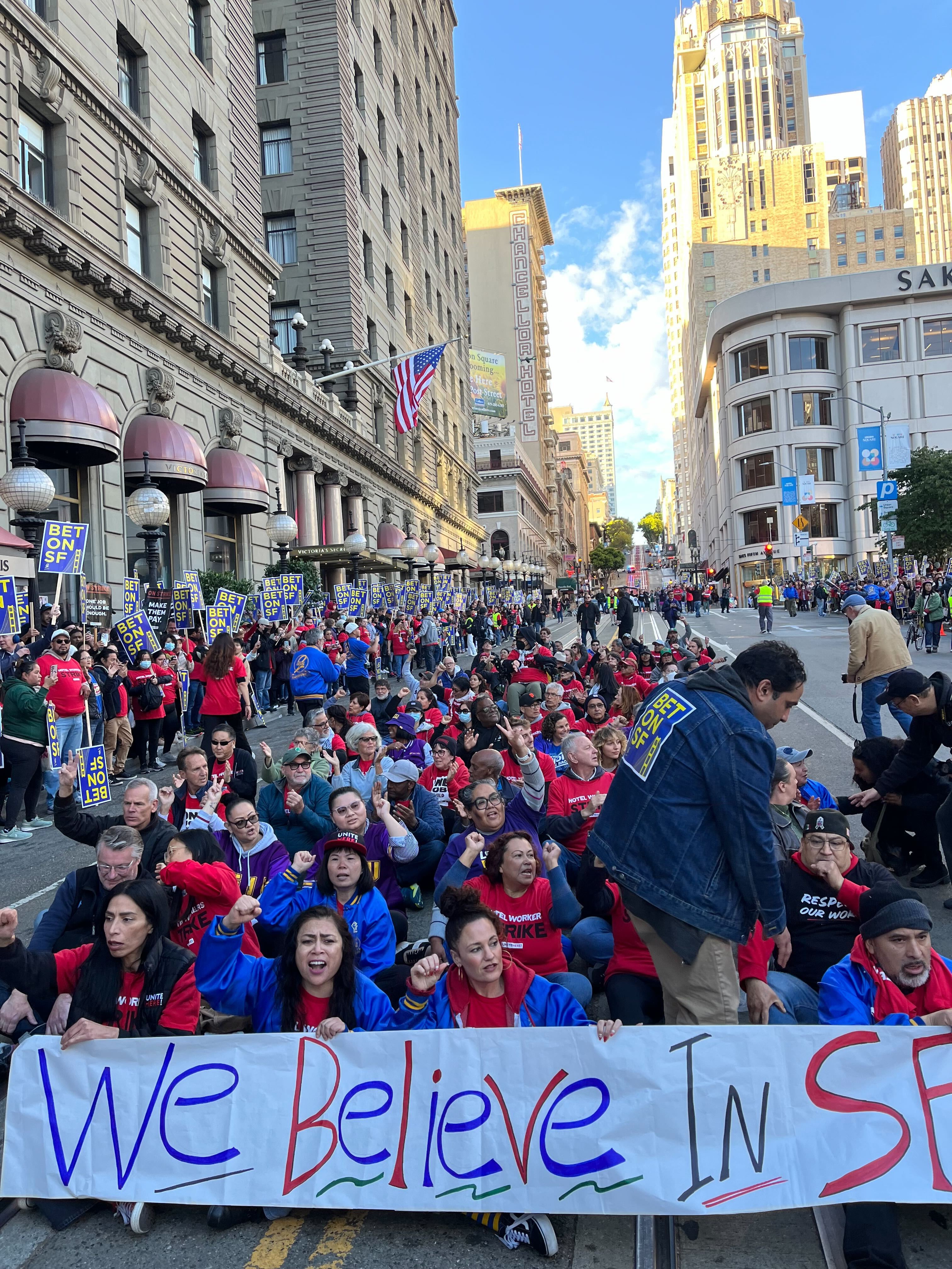 A large group of people sit on a city street, holding signs and a banner that reads &quot;We Believe.&quot; They're gathered near tall buildings, under a clear sky.