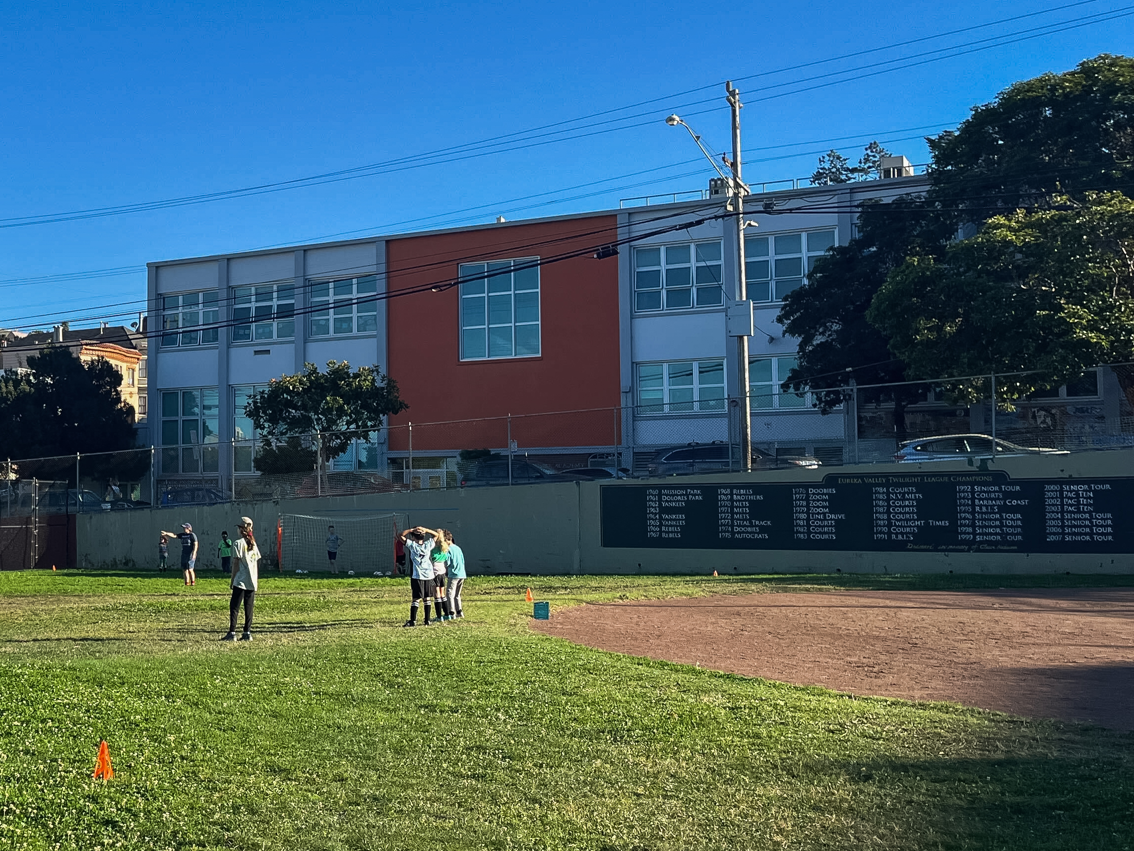 A group of kids in light blue sports jerseys stand on a grassy field near a baseball diamond. A red and white building with trees is in the background.