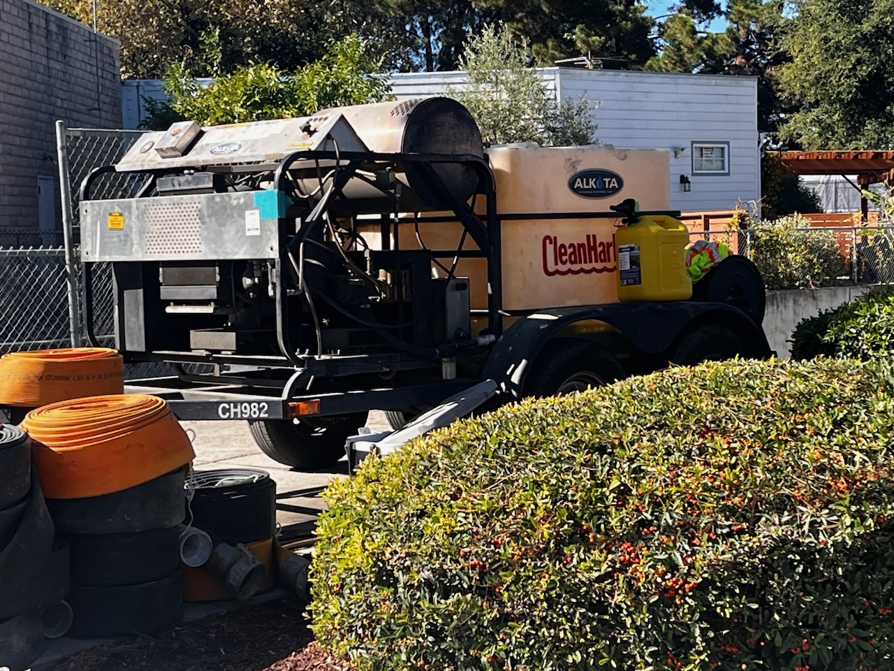 A trailer with industrial equipment and a large &quot;Clean Harbors&quot; tank is parked beside a bush. Coiled orange hoses and pipes are arranged nearby.