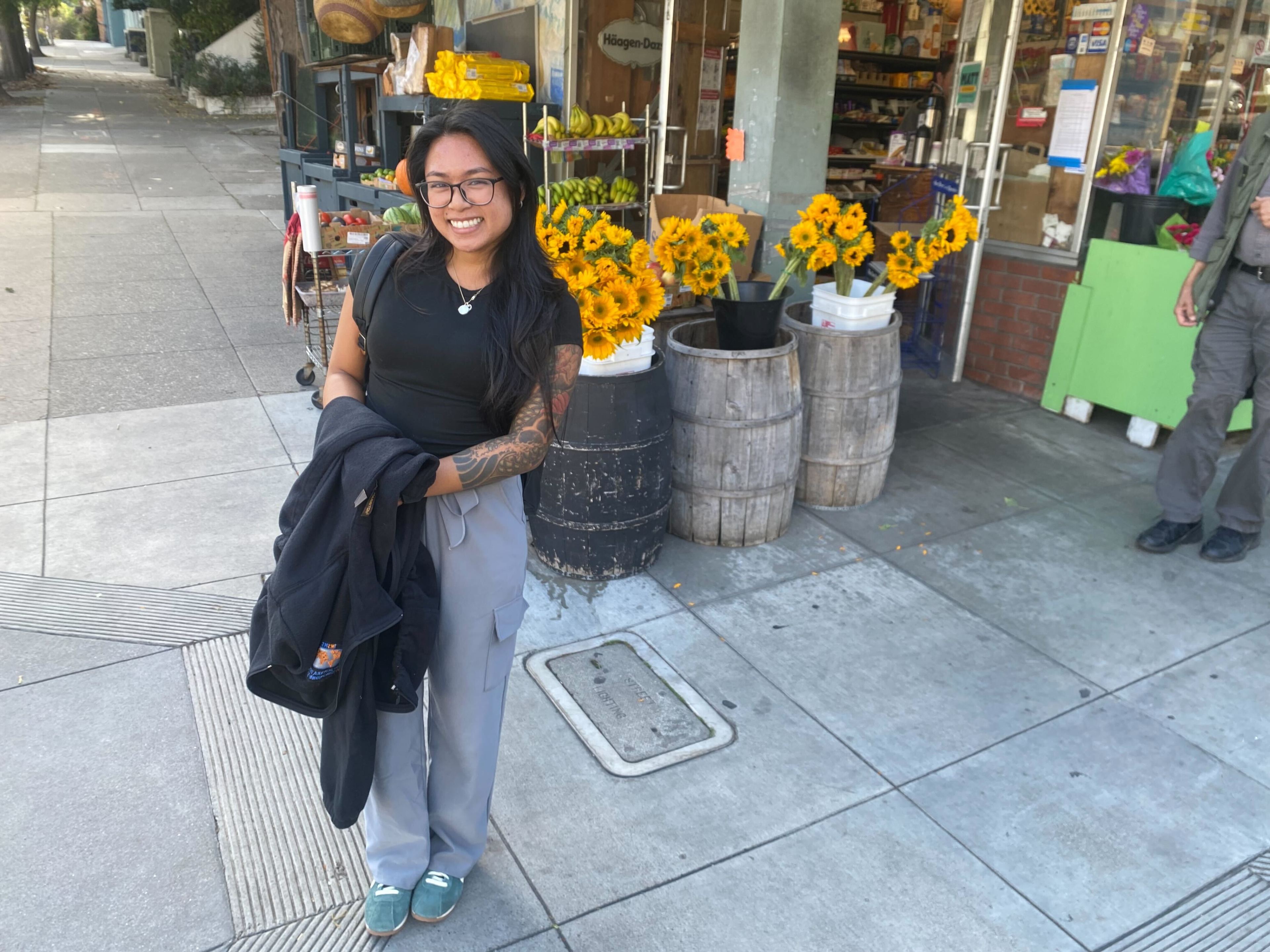 A smiling woman with glasses stands on the street, holding a jacket. She's in front of a store with sunflowers in barrels, and some produce on display.