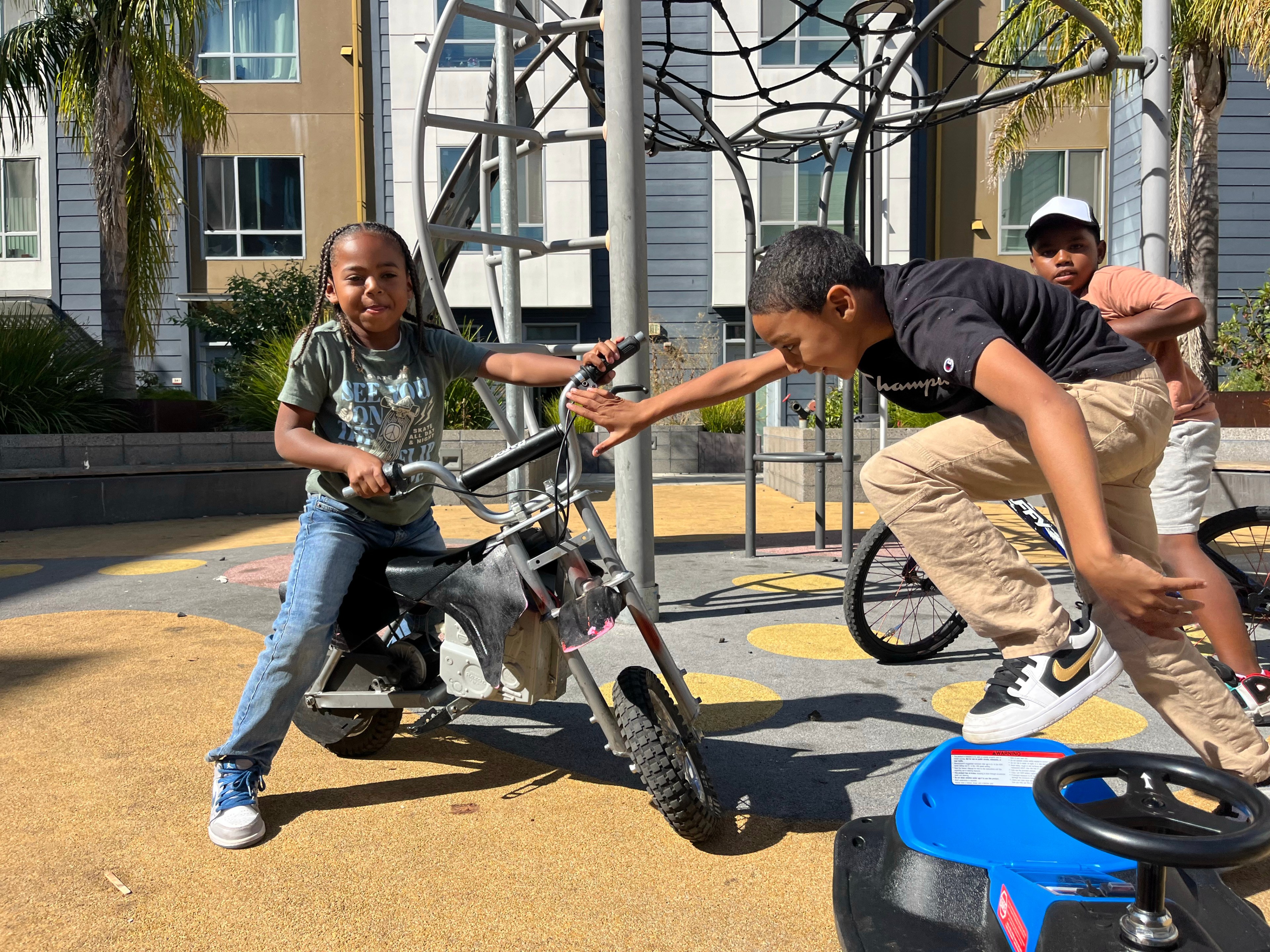 Three children play energetically in a courtyard. One is riding a small bike, another is jumping over a toy car, and the third watches nearby. Buildings surround the area.