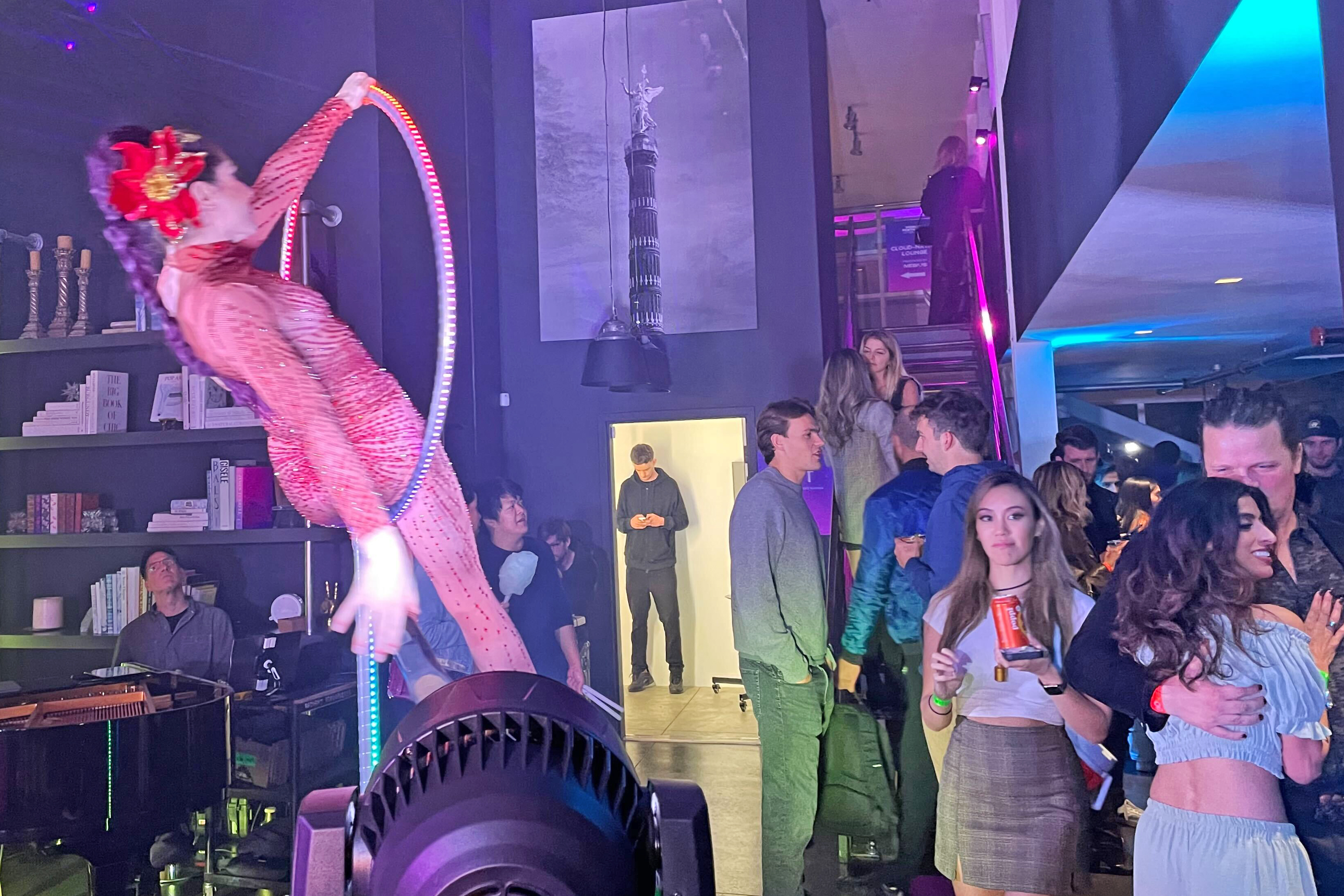 A performer in a red outfit spins in a hoop at a lively party, while guests chat and enjoy drinks in a dimly lit room with colorful lights and a staircase.