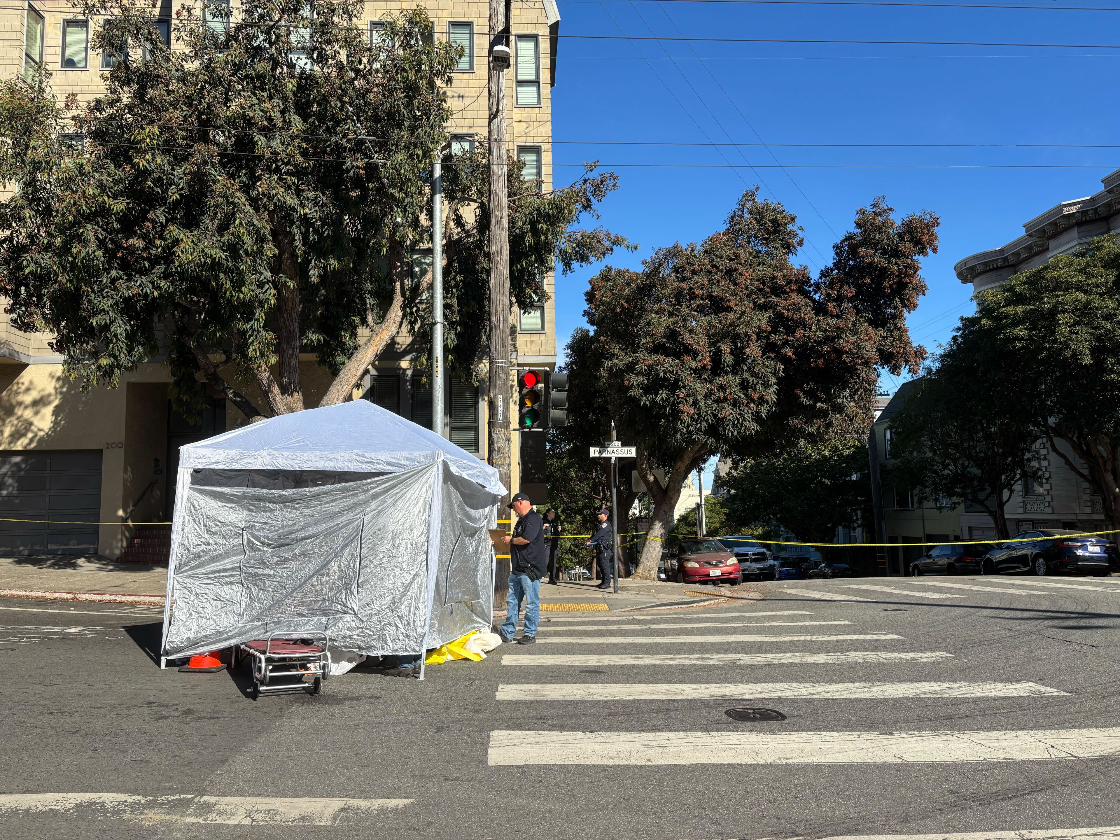 A street scene shows a white tent set up at an intersection, partially blocking the crosswalk. A person is standing nearby, and trees line the sidewalks.