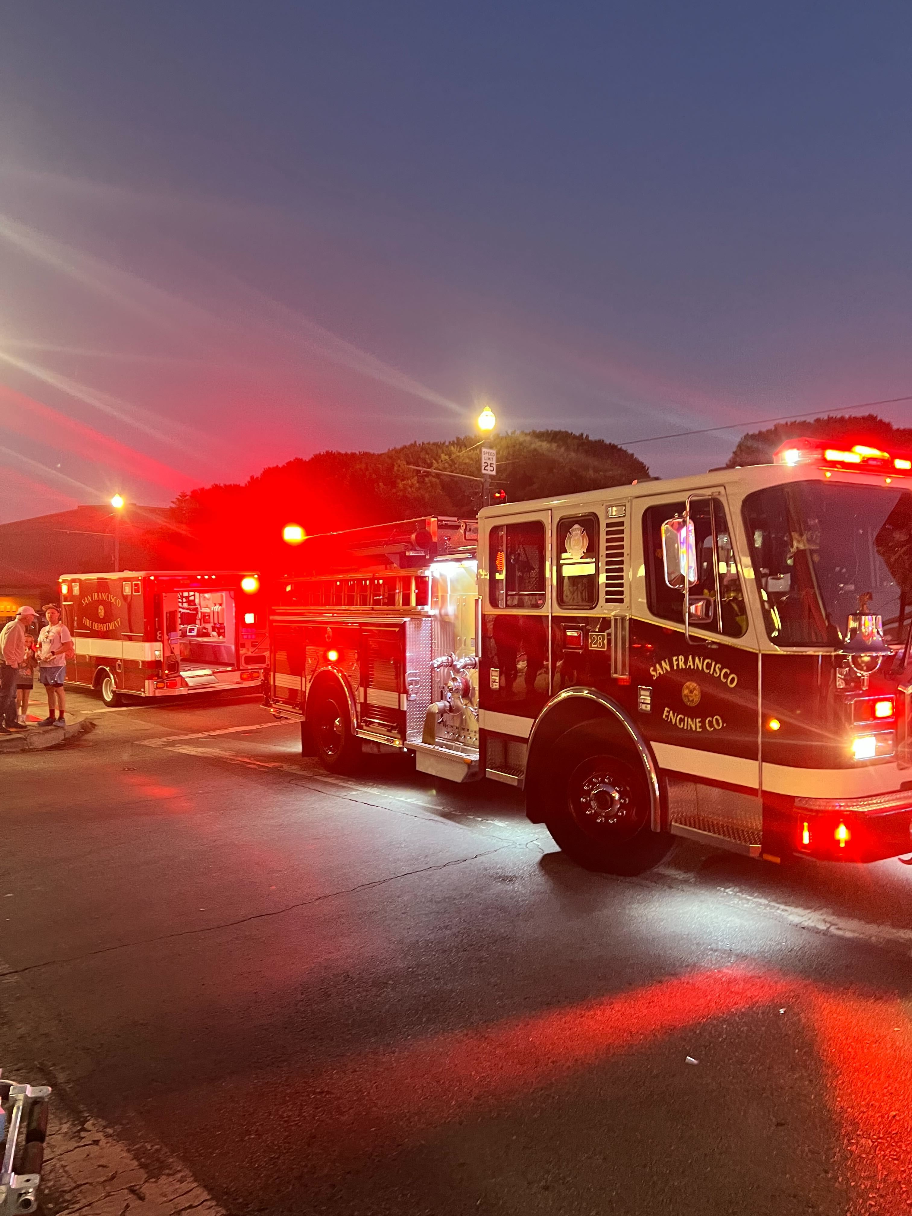 A fire engine with flashing red lights is parked on a street at dusk. The scene includes another emergency vehicle and a few people standing nearby.