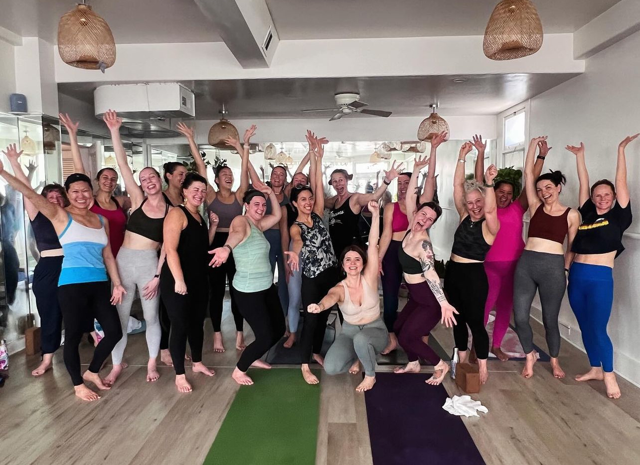 A joyful group of people poses energetically in a yoga studio, with yoga mats on the floor. They are smiling and raising their arms, creating a lively atmosphere.