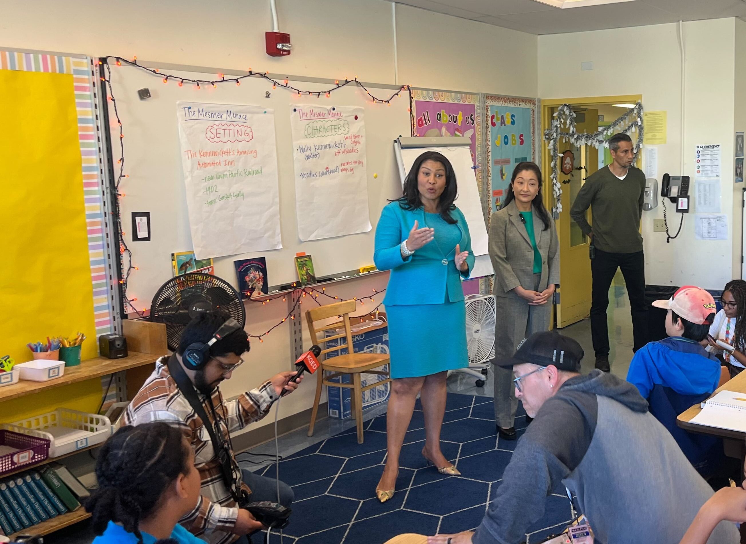 A woman in a blue outfit speaks to a classroom full of attentive students. A cameraman and another adult are present. The room is decorated with educational materials.