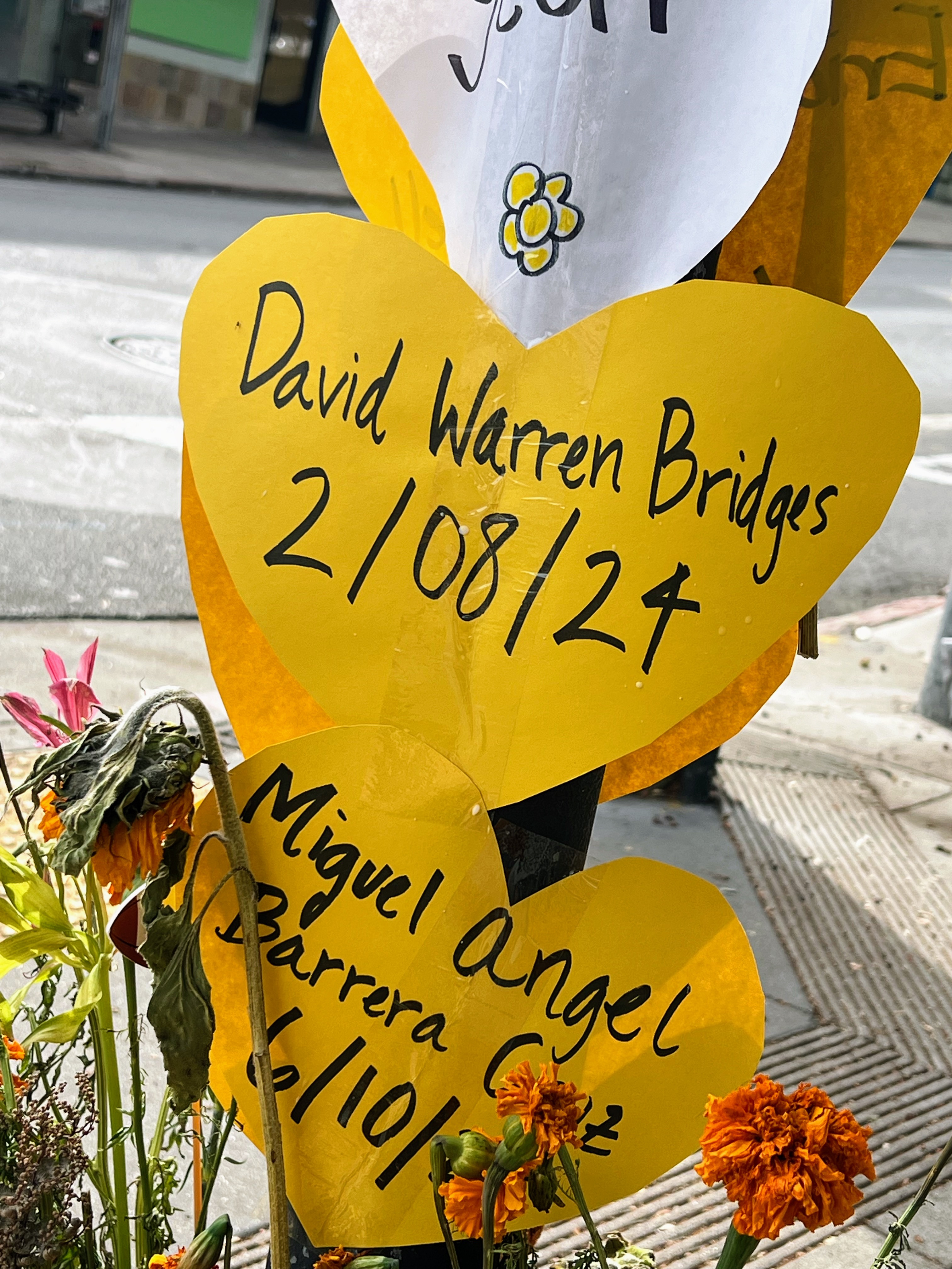 Yellow heart-shaped signs on a pole with handwritten names and dates. Flowers are placed below, partially wilted and vibrant marigolds.