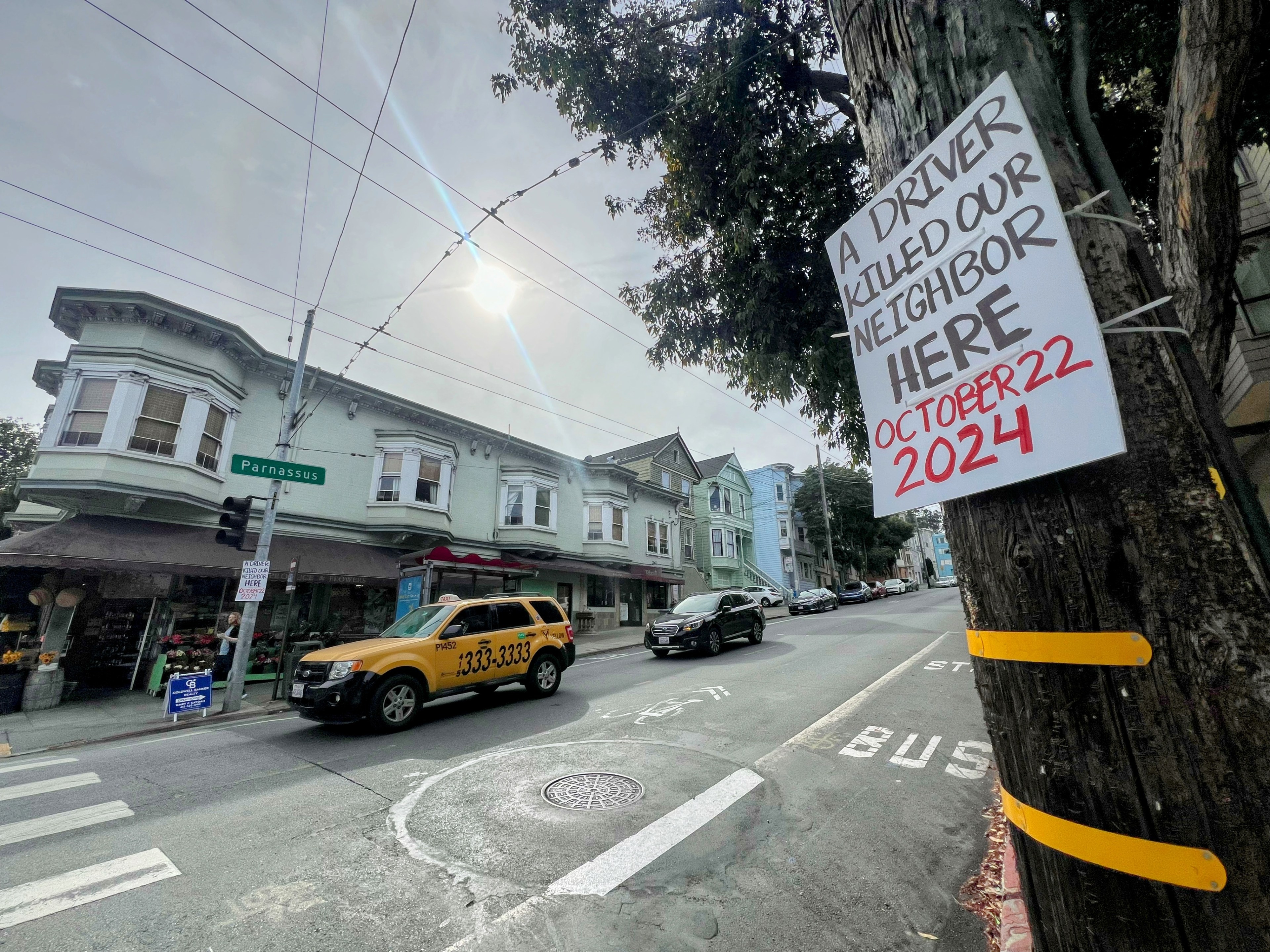 A street corner features a pole with a sign reading: &quot;A driver killed our neighbor here, October 22, 2024.&quot; A yellow taxi is parked by shops.