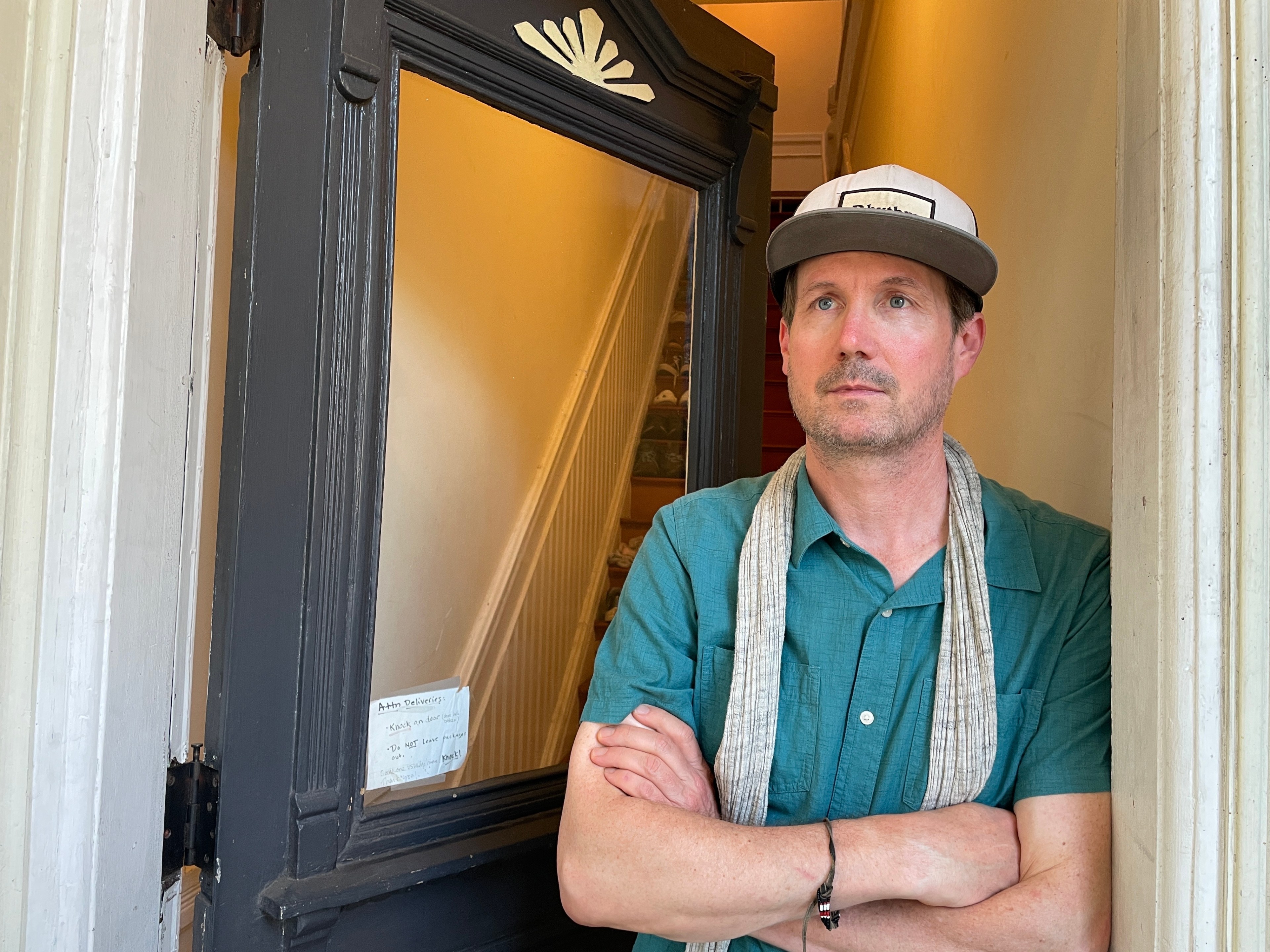 A man in a blue shirt and hat stands in a doorway with crossed arms, a beige scarf around his neck, and an interior stairway visible behind him.