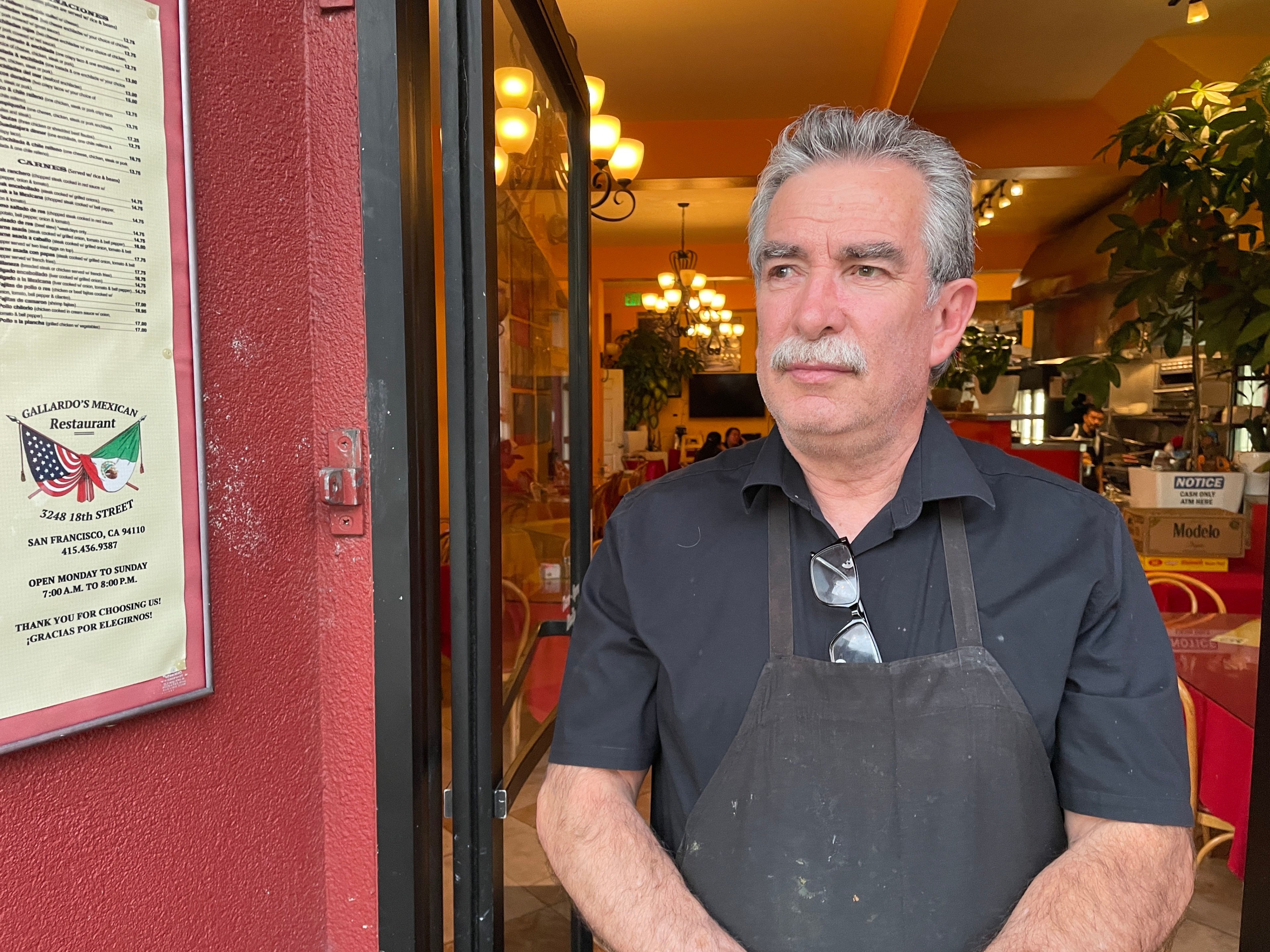 A man with gray hair and a mustache stands at the entrance of a Mexican restaurant, wearing a black shirt and apron. A menu is displayed on the wall beside him.