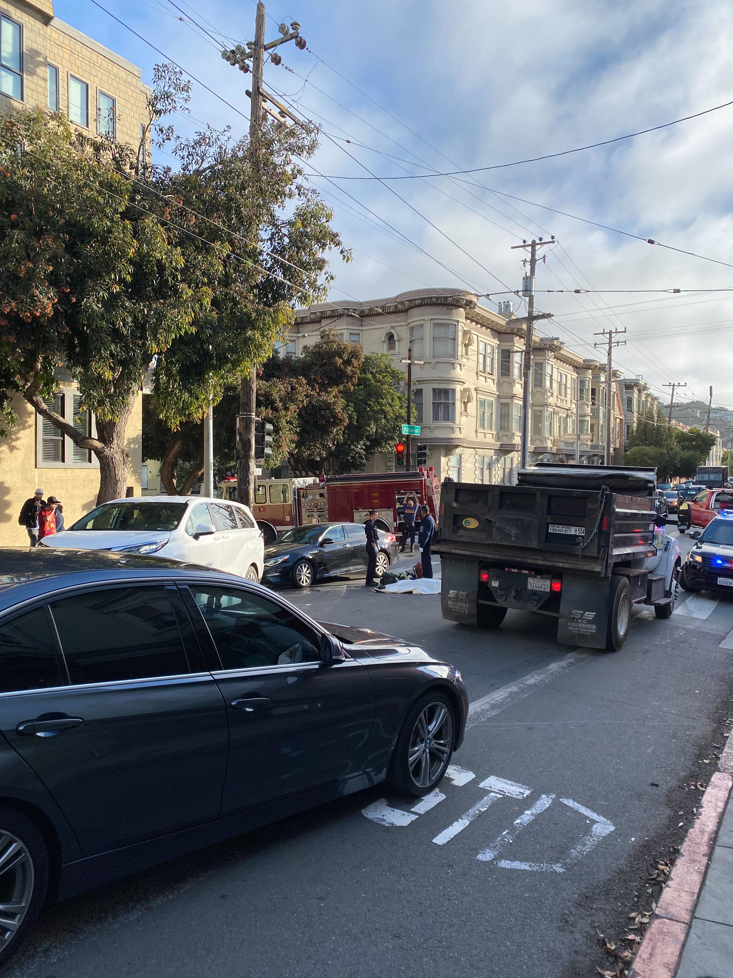 The image shows a busy street scene with multiple cars, a fire truck, and emergency responders attending to an incident. Nearby buildings and trees are visible.