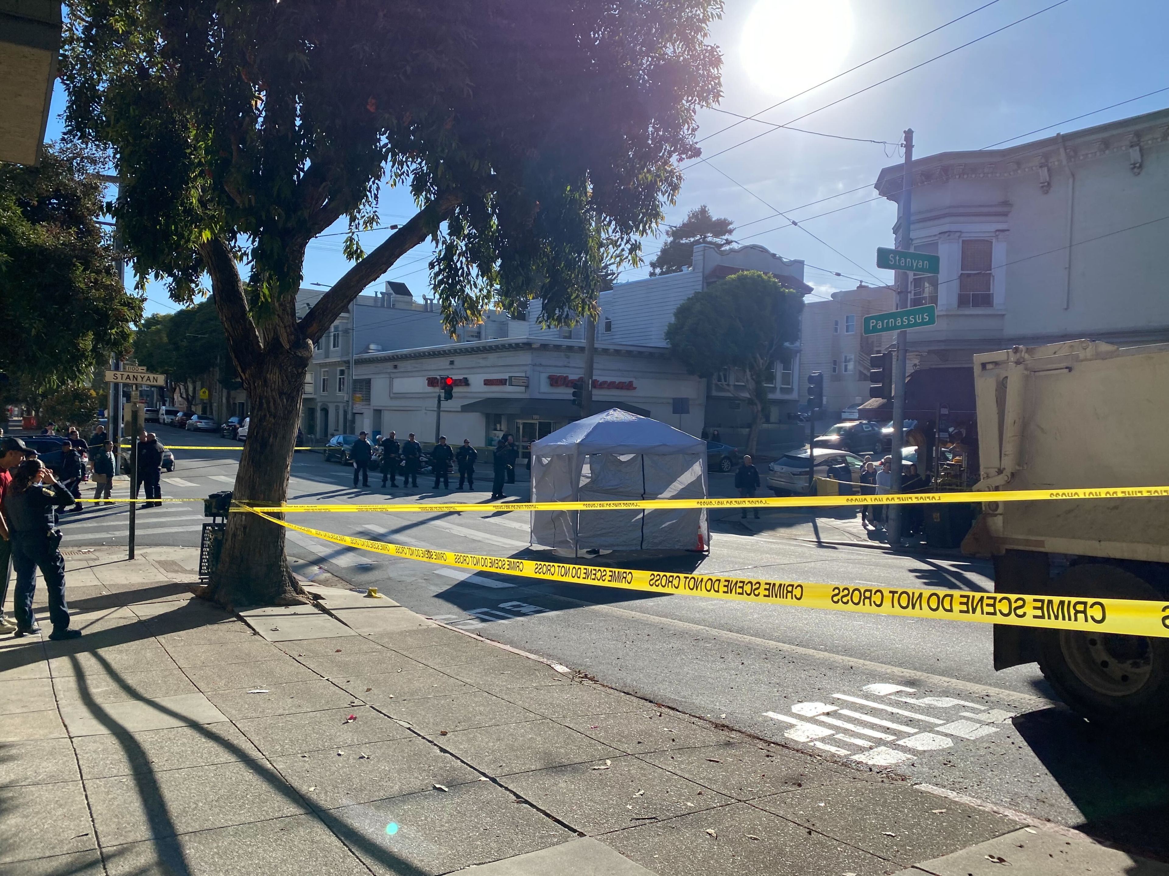 A police scene is cordoned off with yellow tape at the corner of Stanyan and Parnassus streets. Officers and a small tent are present under a clear, sunny sky.