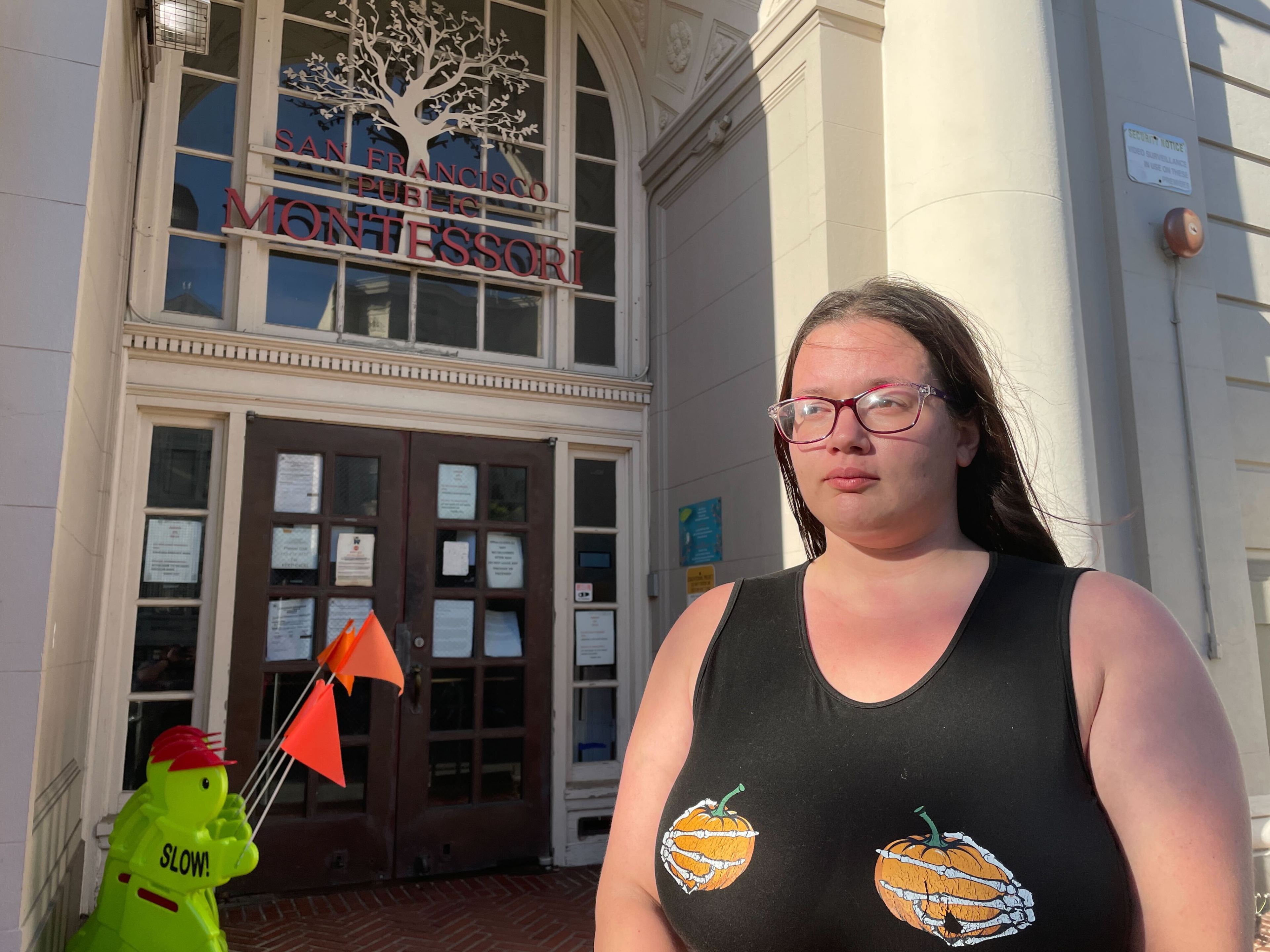 A person stands outside a building labeled &quot;San Francisco Public Montessori.&quot; They're wearing glasses and a black tank top with pumpkin designs.