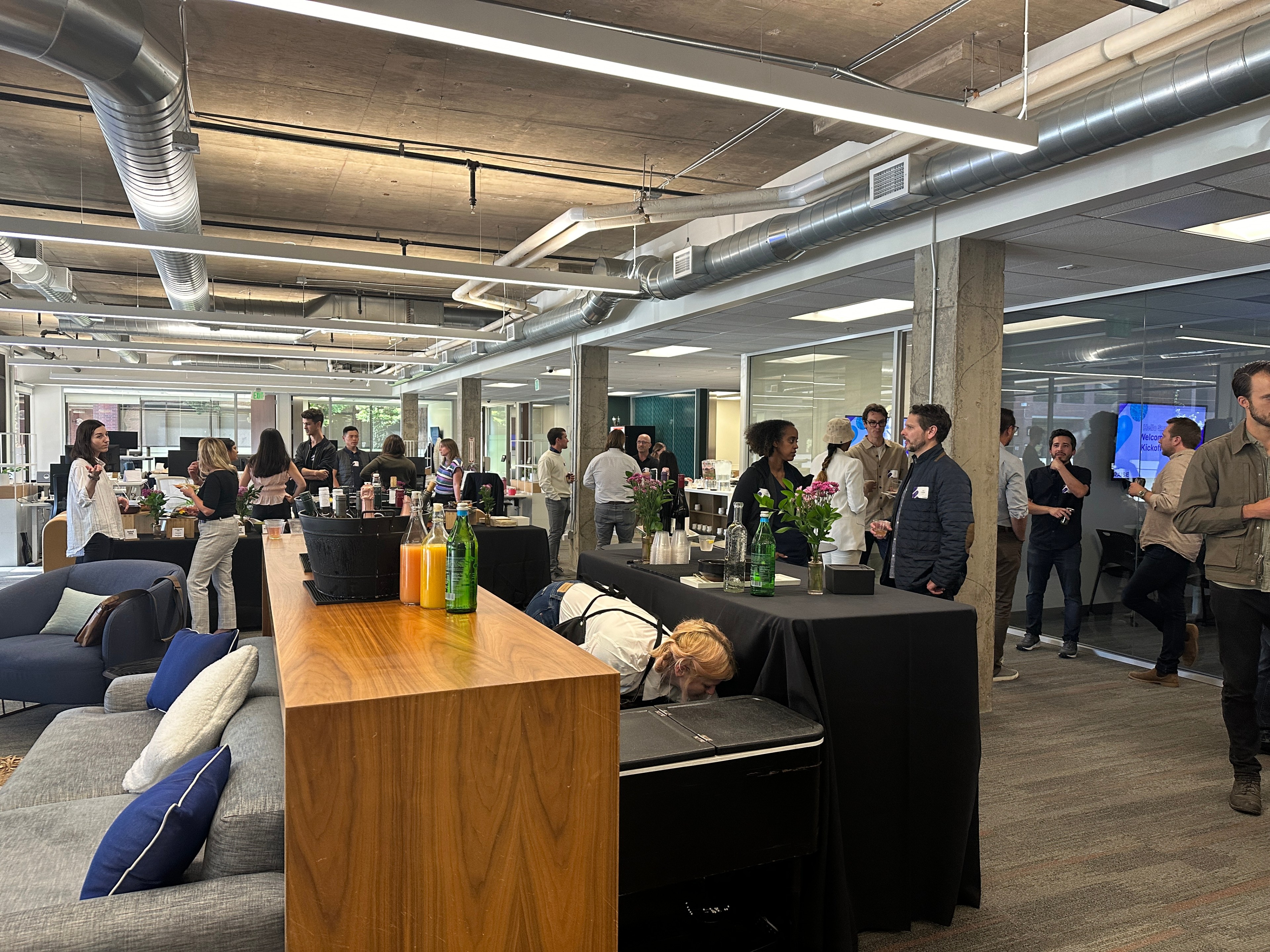 The image shows a busy office gathering with people socializing around tables with drinks. The setting is modern, with exposed ductwork and comfortable seating.