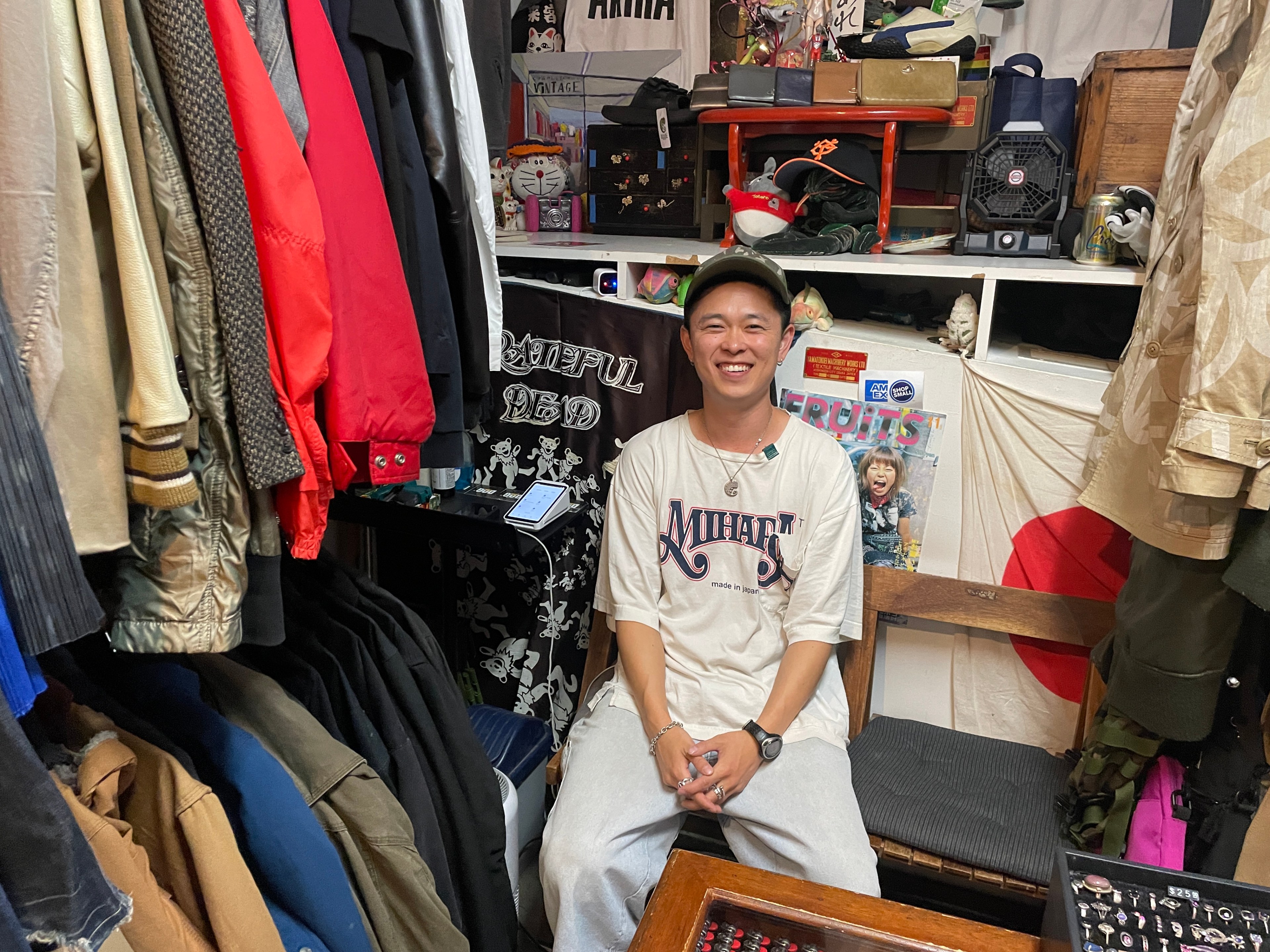 A person is sitting and smiling in a cluttered room filled with clothes, posters, and various items. He wears a baseball cap and a white graphic shirt.