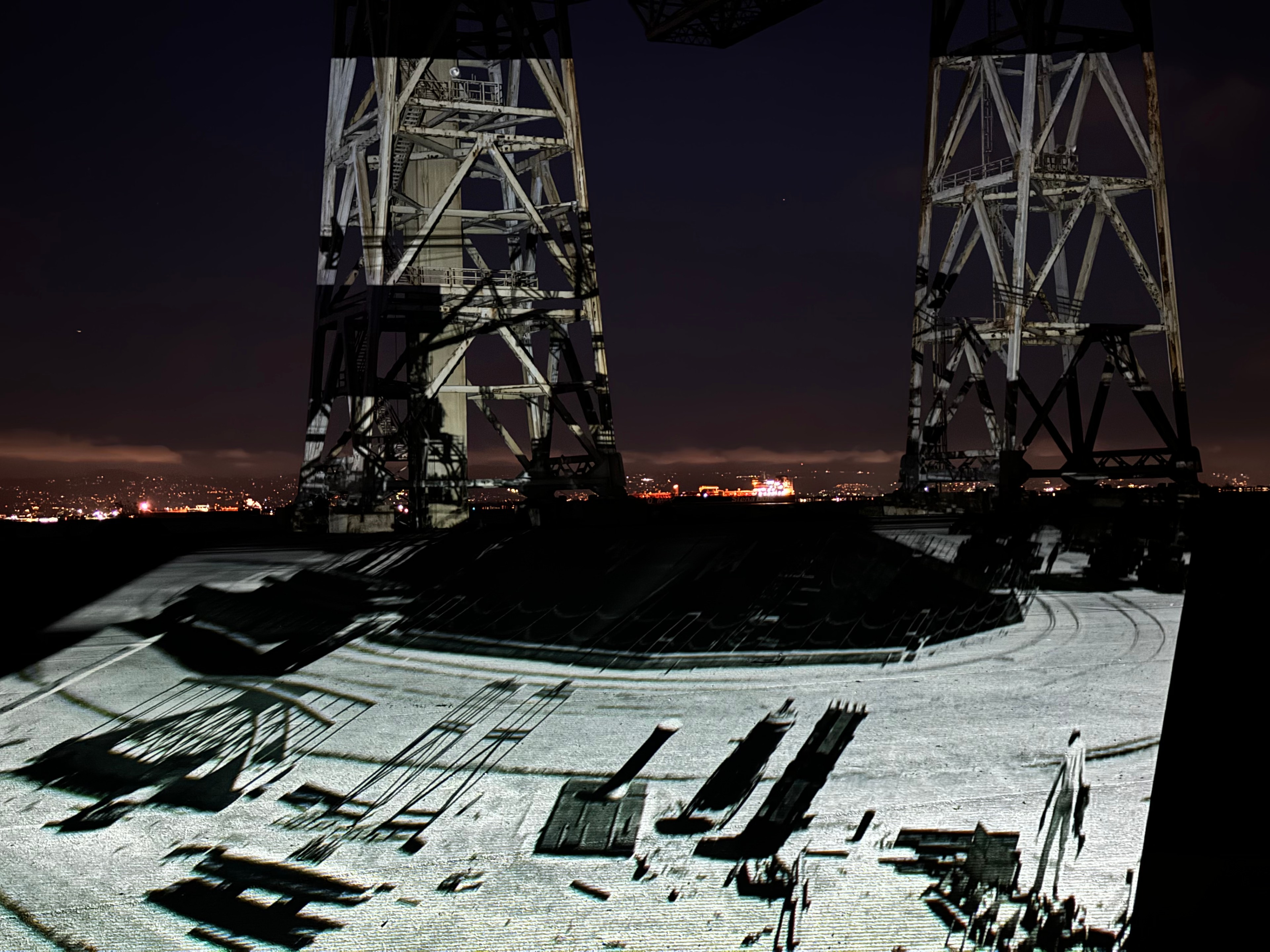 A night scene with a large metal structure and projected images on the ground. In the distance, city lights and a ship are visible against a dark sky.