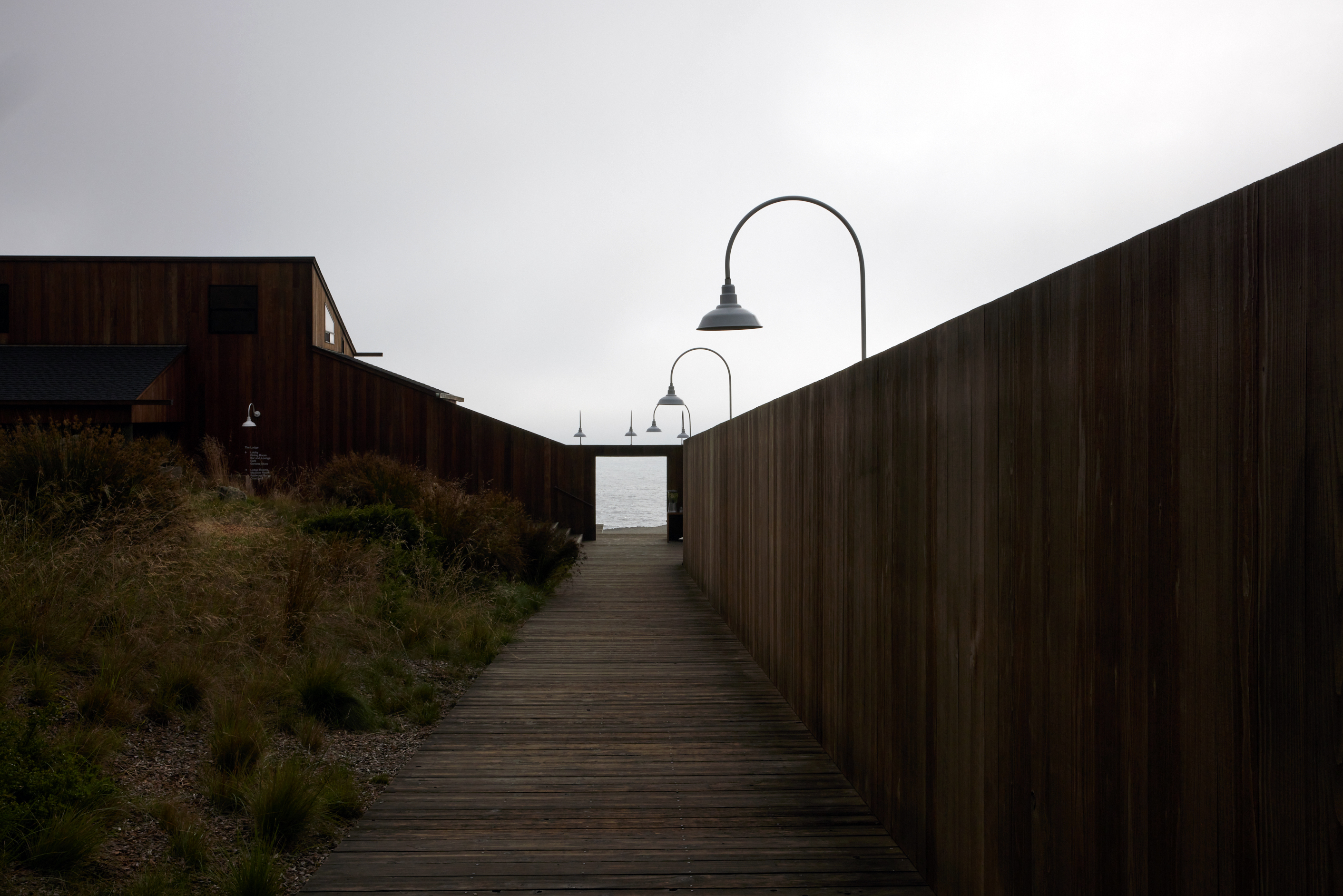 A wooden pathway leads to the sea, flanked by tall wooden walls and lit by street lamps under a cloudy sky. Grassy vegetation borders the path on one side.