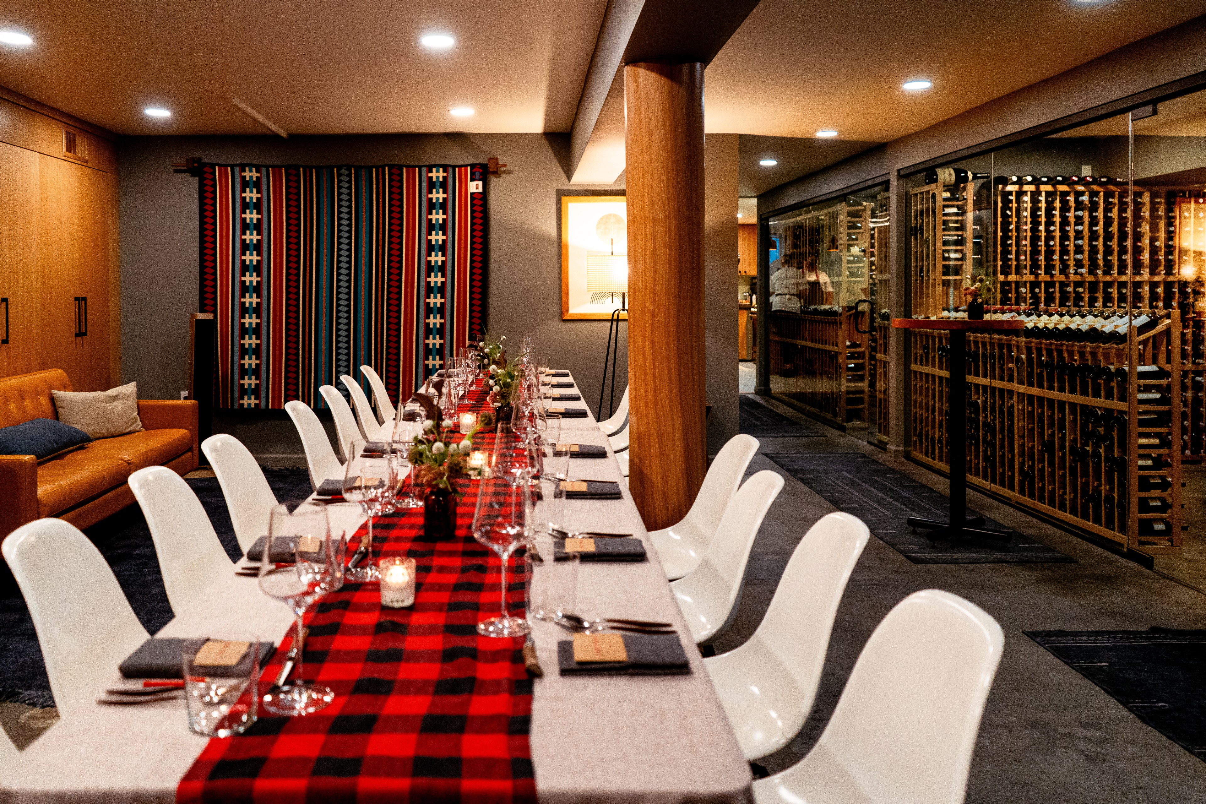 A long dining table is elegantly set with wine glasses, candles, and a red plaid runner. It is surrounded by white chairs, with a cozy sofa and a wine cellar nearby.