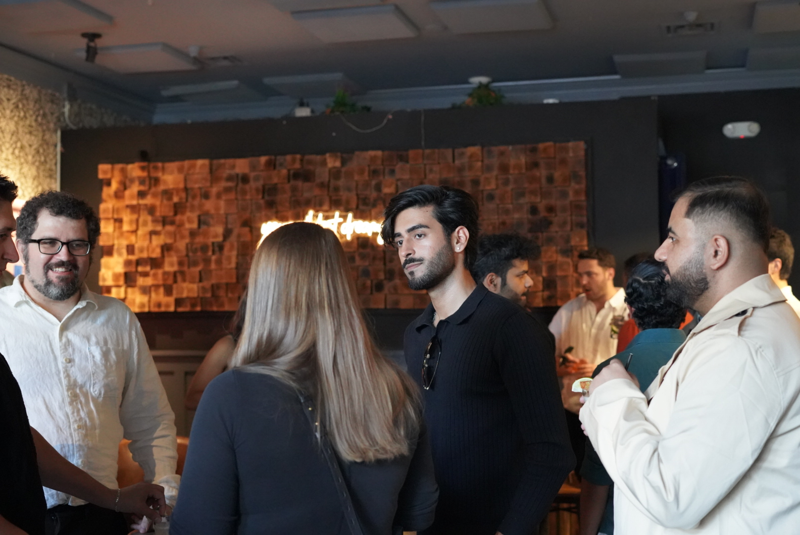 A group of people are chatting in a warmly lit indoor space with a wooden tiled wall and neon sign in the background. They appear to be at a casual gathering.