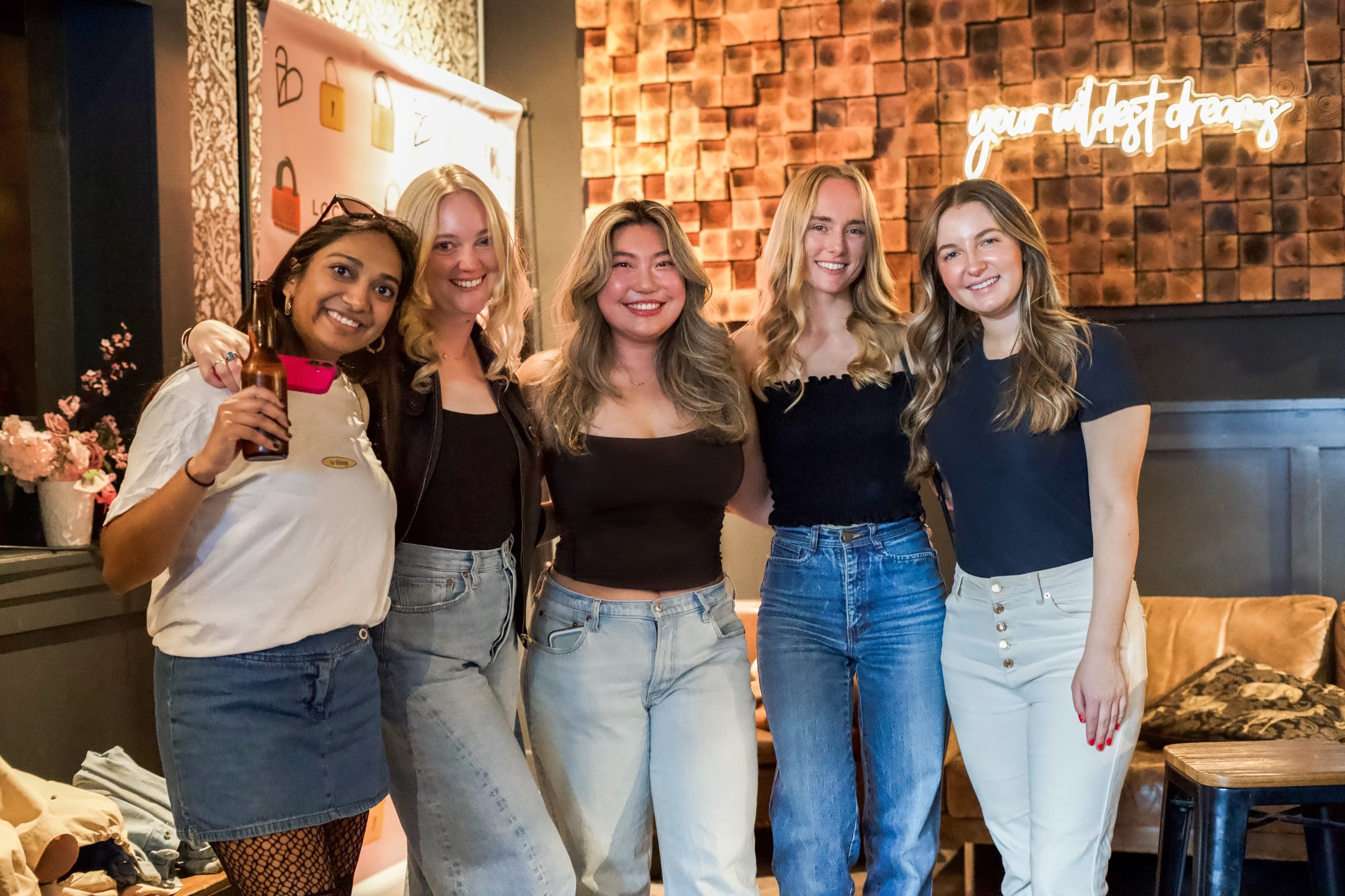 Five smiling women stand close together, posing indoors with cozy decor. One holds a drink, and there's a neon sign in the background.