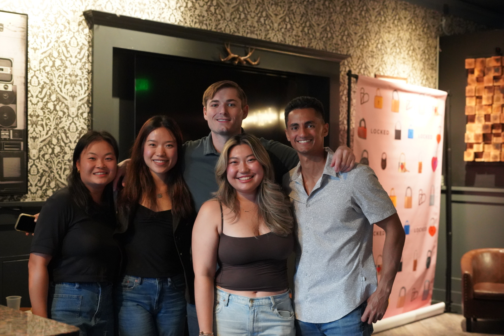 Five people are standing together indoors, smiling at the camera. There's patterned wallpaper and a banner with locks behind them. They appear to be at an event.