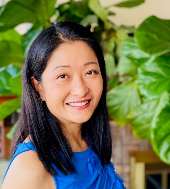 A woman with long black hair is smiling while wearing a blue top. She stands in front of large green leaves, creating a vibrant, cheerful background.