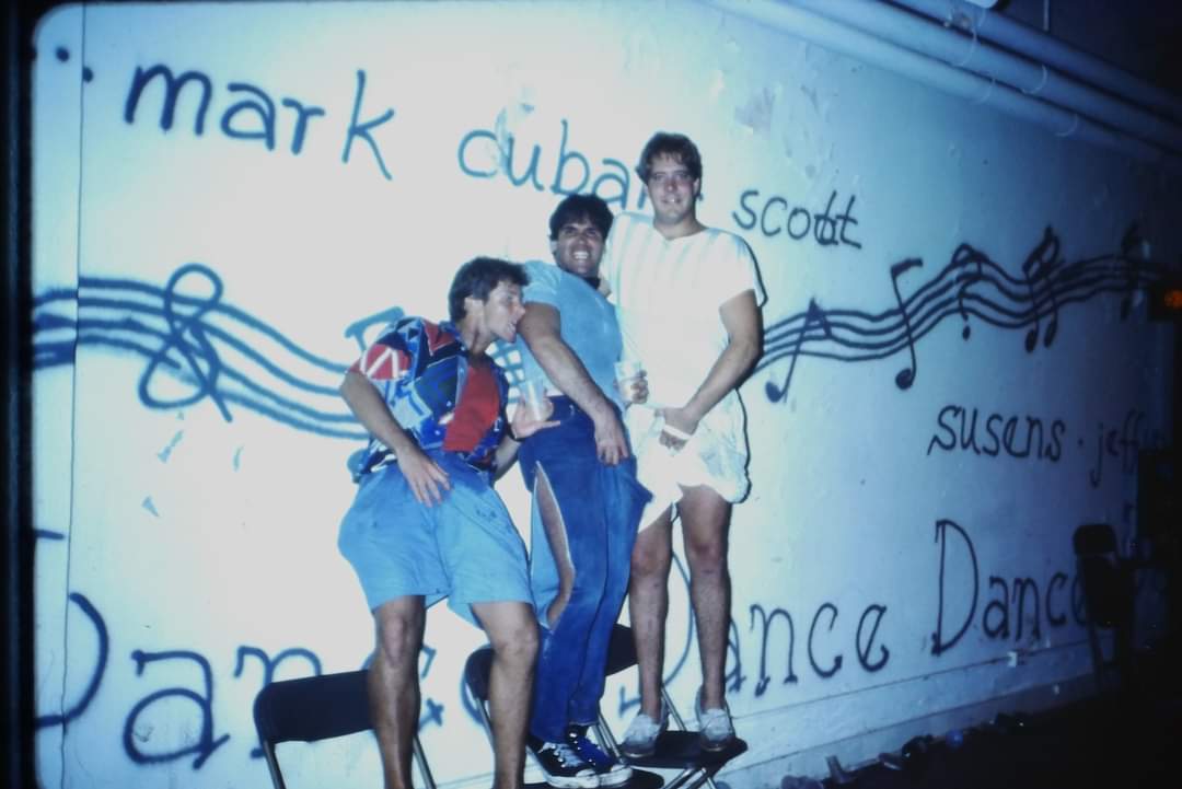 Three men pose playfully against a wall with musical notes and names painted on it. They're standing closely, with casual outfits, in a dimly lit room.