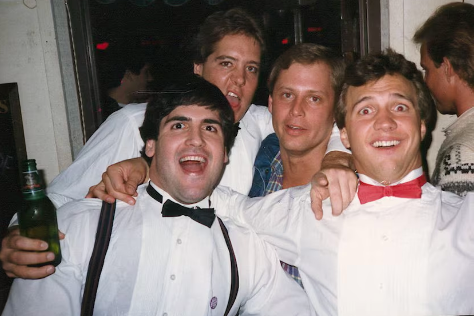 Four men are smiling and posing for a group photo. Two wear black bow ties, one wears a red bow tie, and there is a drink in hand. They seem happy and festive.