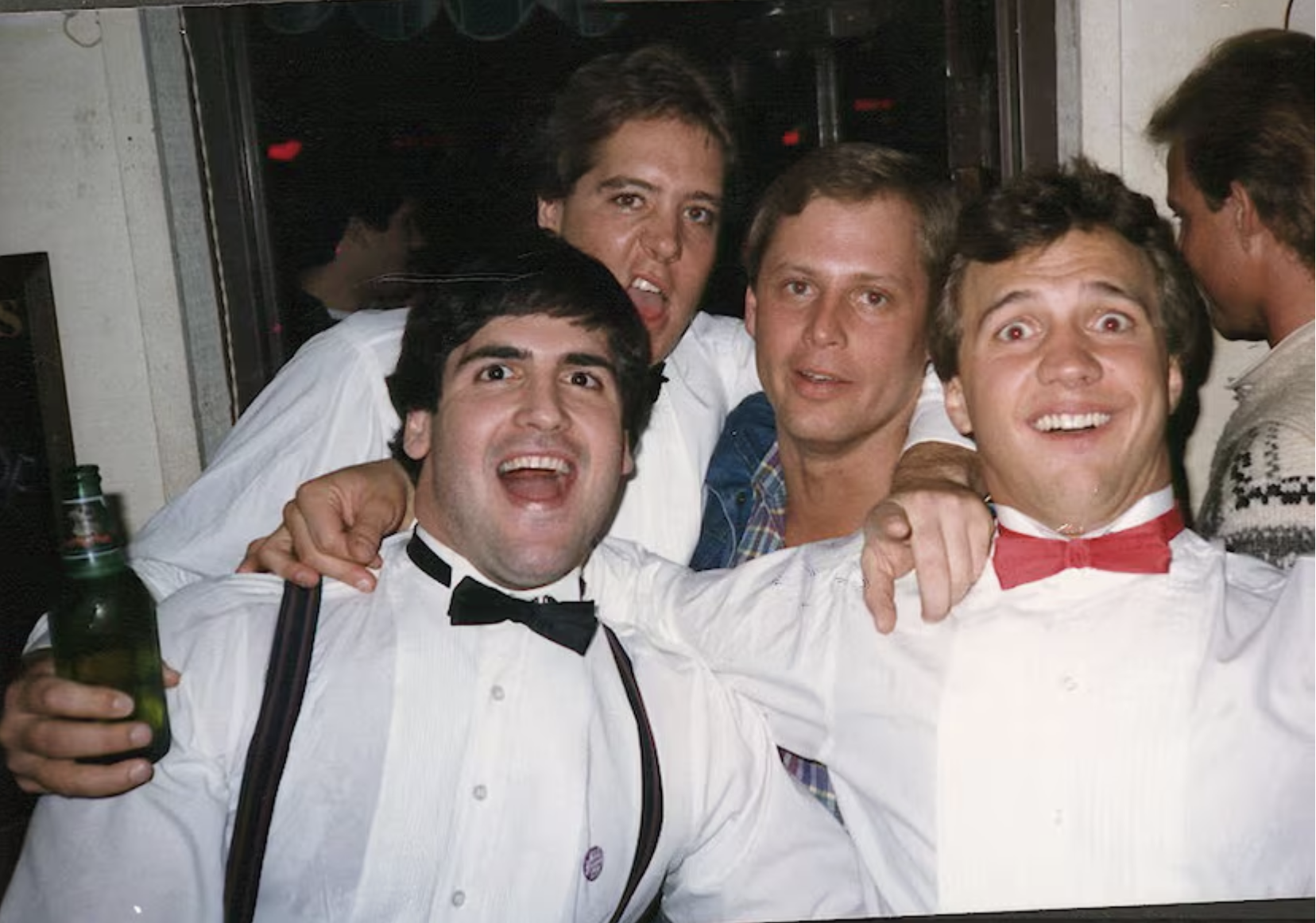 Four men are smiling and posing for a group photo. Two wear black bow ties, one wears a red bow tie, and there is a drink in hand. They seem happy and festive.