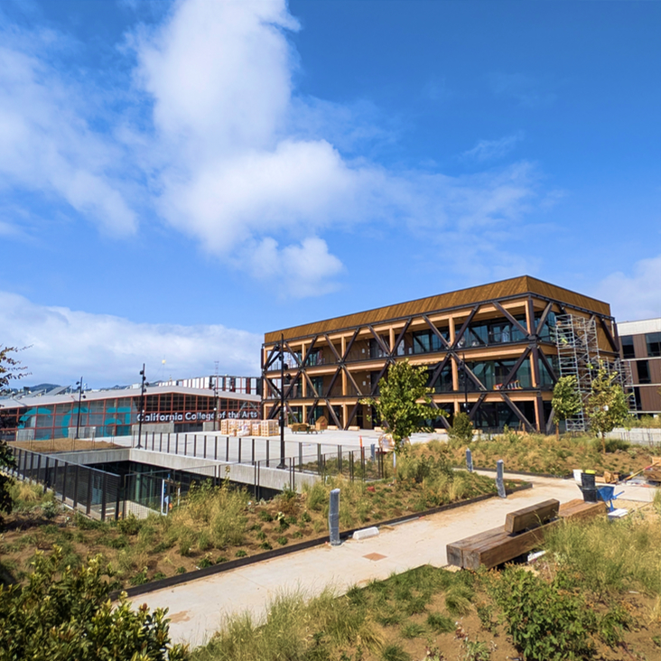 The image shows a modern building with wooden exteriors and large windows, surrounded by greenery. A sign reads &quot;California College of the Arts&quot; under a clear blue sky.