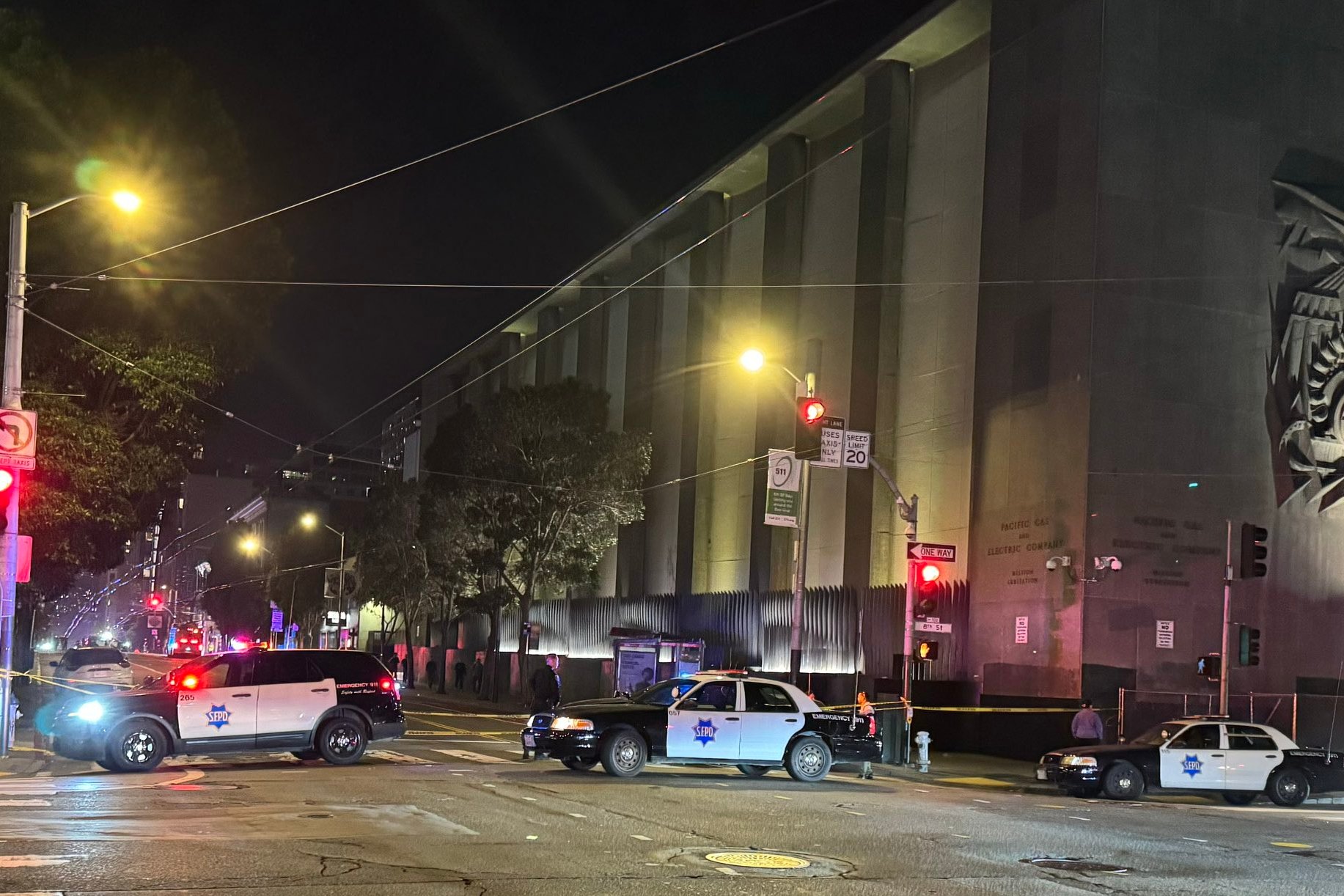 Three police cars block a street at night near a large building with sculpted art on the wall. Bright streetlights illuminate the scene.