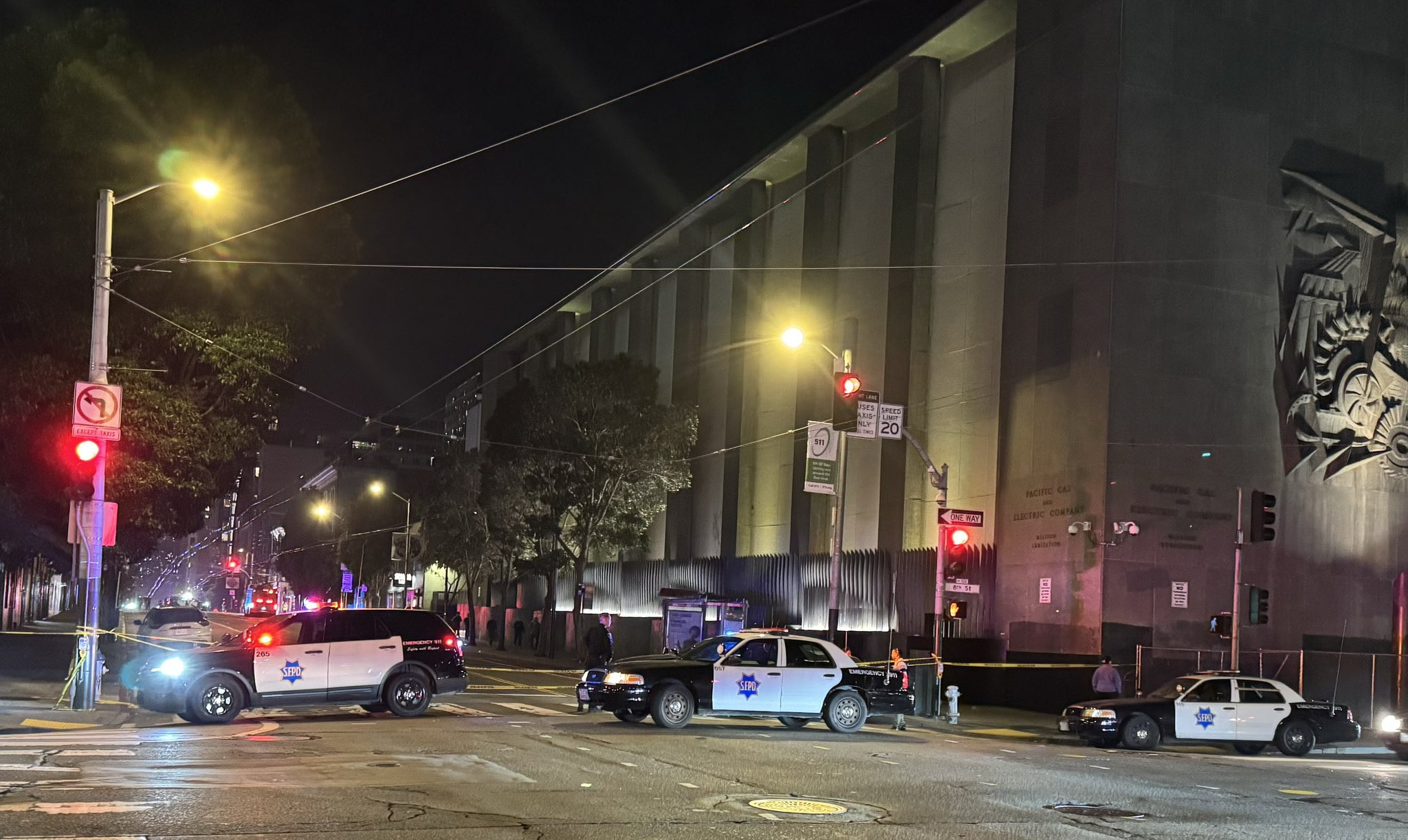 Three police cars block a street at night near a large building with sculpted art on the wall. Bright streetlights illuminate the scene.