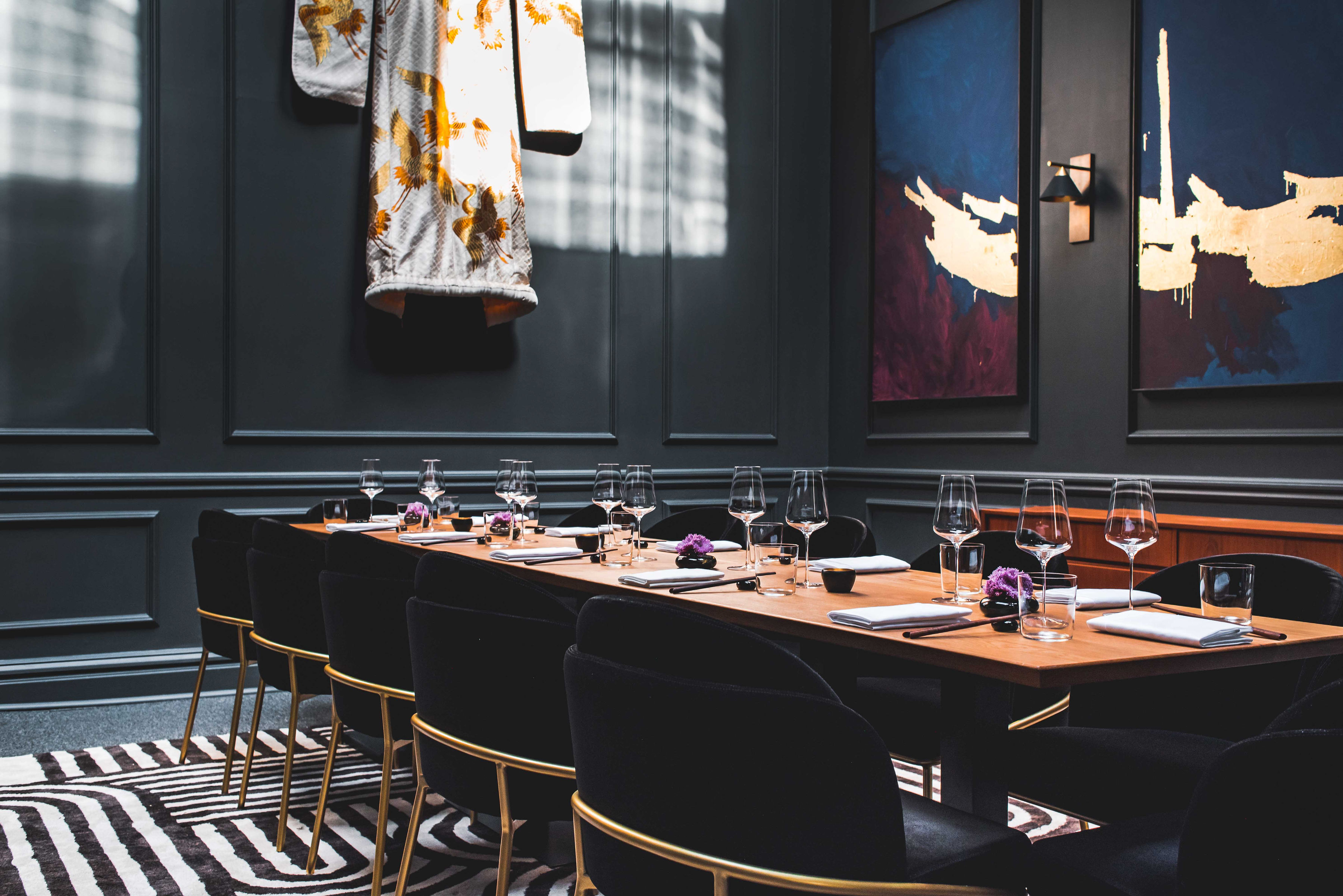 A stylish dining room with a long wooden table set with glasses, napkins, and small purple flowers. Dark walls feature abstract art and a wall-hanging kimono.