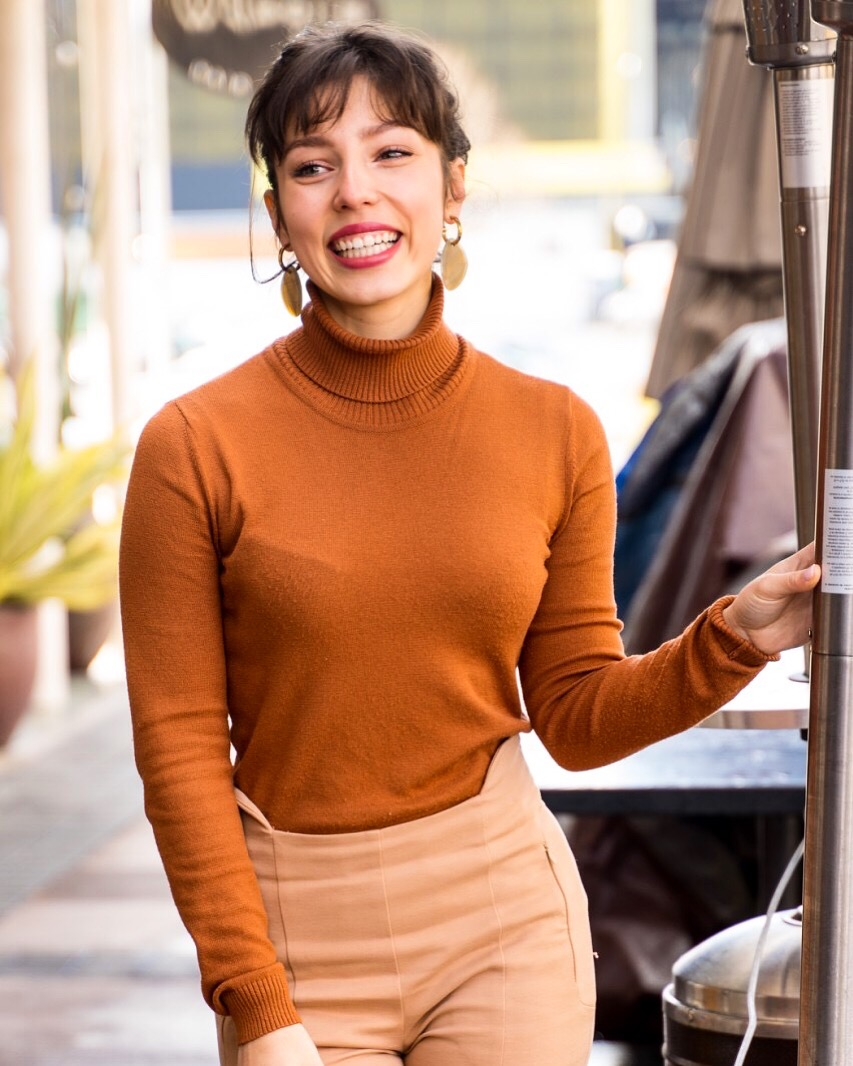 A smiling woman wearing a rust-colored turtleneck and beige pants stands outdoors, holding a vertical pole. She has short hair and large hoop earrings.