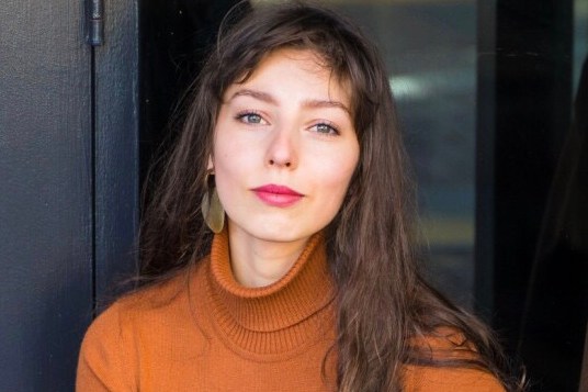 The image shows a woman with long, wavy brown hair, wearing a rust-colored turtleneck sweater and large gold earrings. She is seated in front of a dark background.