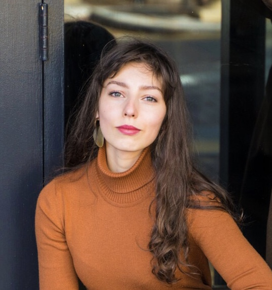 The image shows a woman with long, wavy brown hair, wearing a rust-colored turtleneck sweater and large gold earrings. She is seated in front of a dark background.