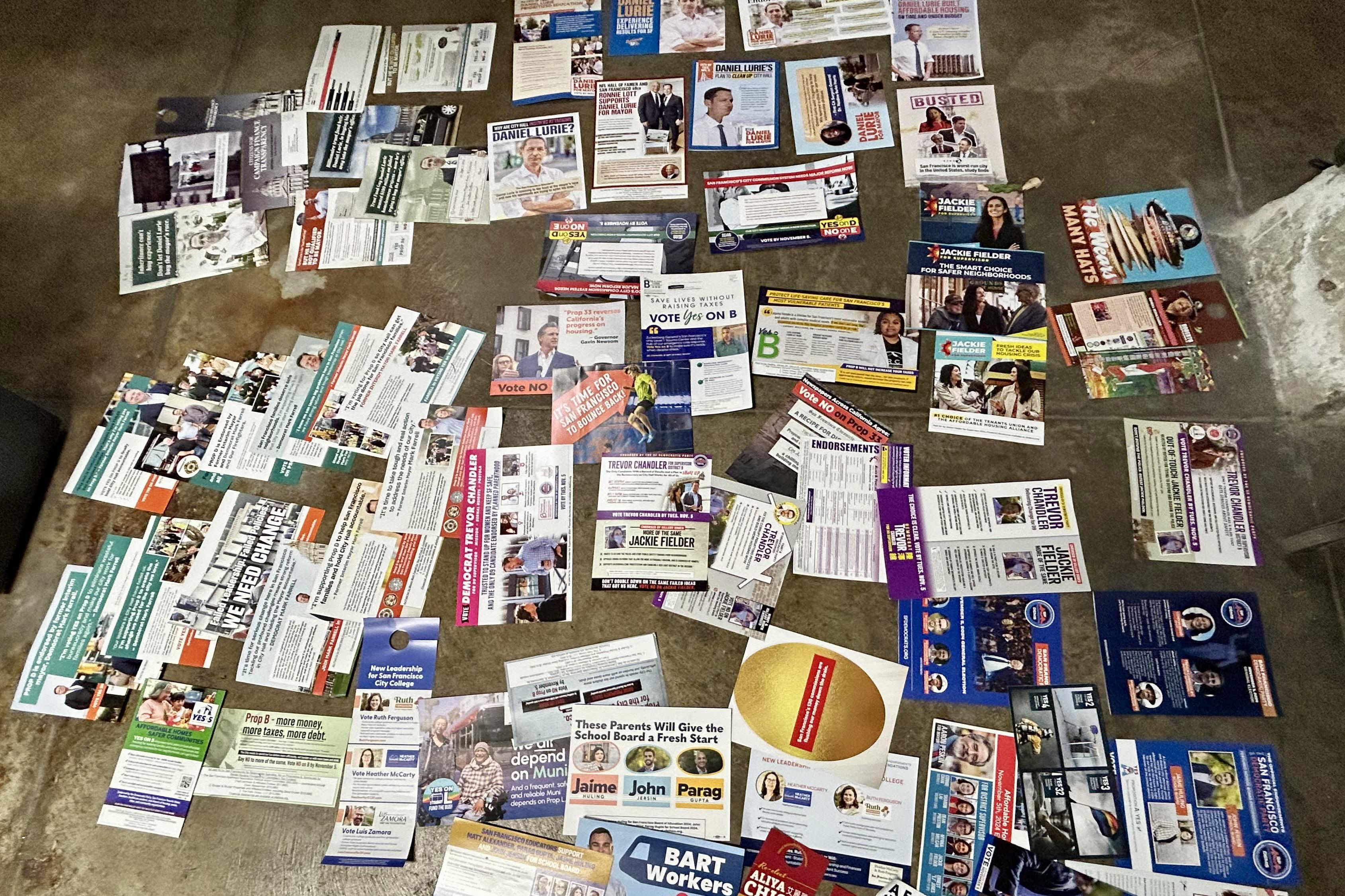 The image shows numerous political flyers scattered on a concrete floor, featuring various candidates, propositions, and colorful campaign materials.