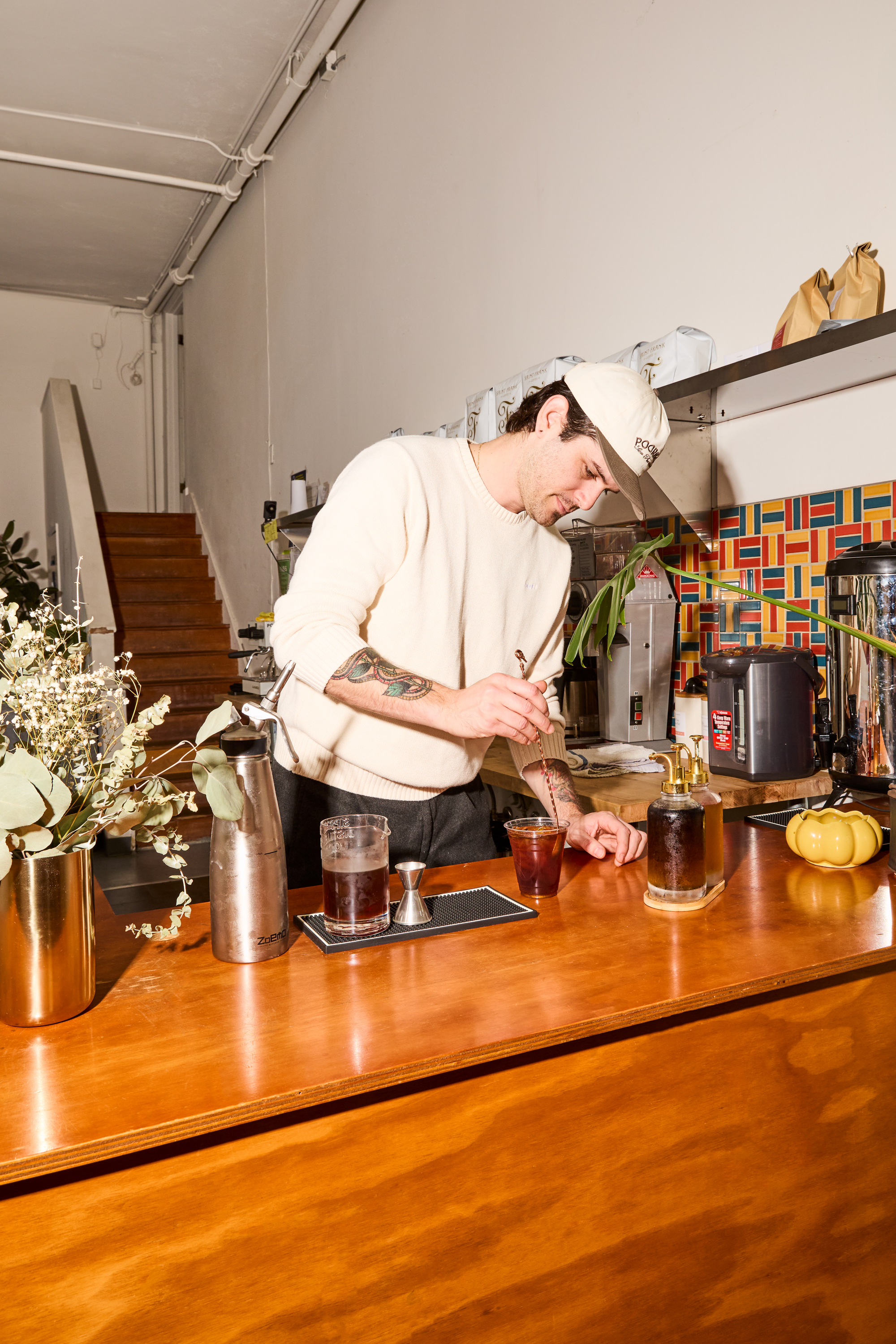 A person in a cream sweater and cap prepares a drink at a wooden bar, surrounded by coffee equipment, flowers, and a small pumpkin decoration.