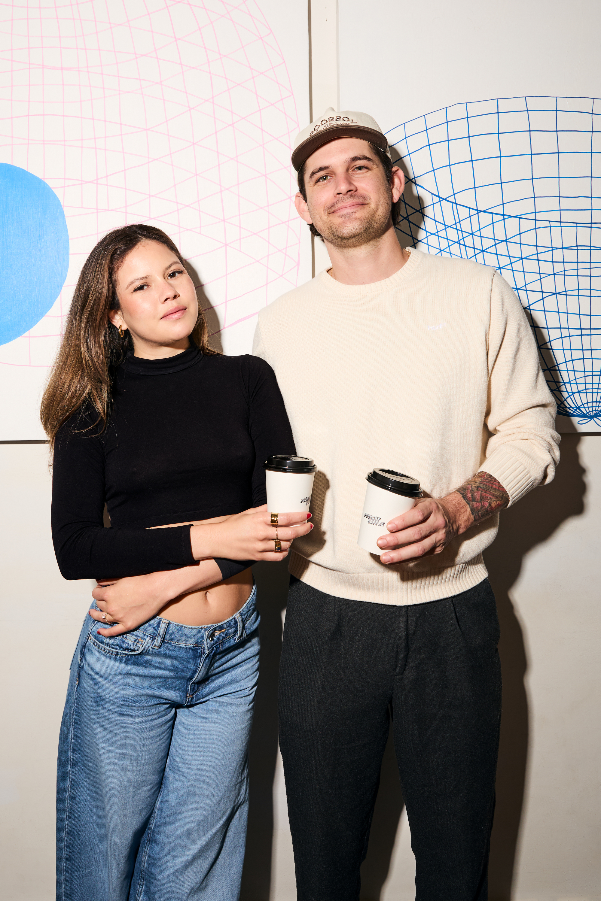 A man and woman stand smiling, holding coffee cups. The woman wears a black top and jeans, while the man wears a cream sweater and cap. Geometric art is behind them.