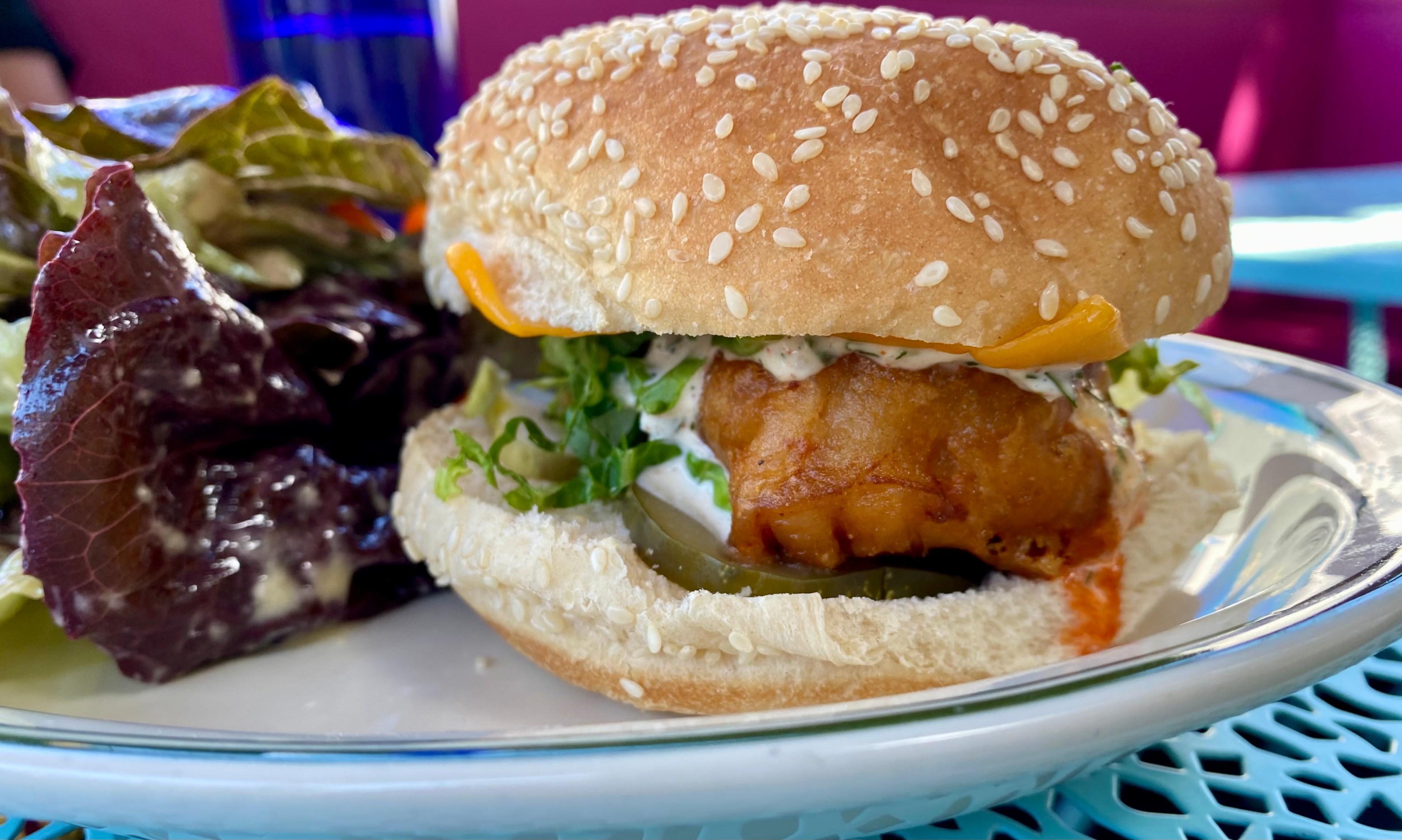 A sesame seed bun burger with crispy filling, lettuce, pickles, sauce, and a cheese slice is on a plate next to some mixed leafy greens.